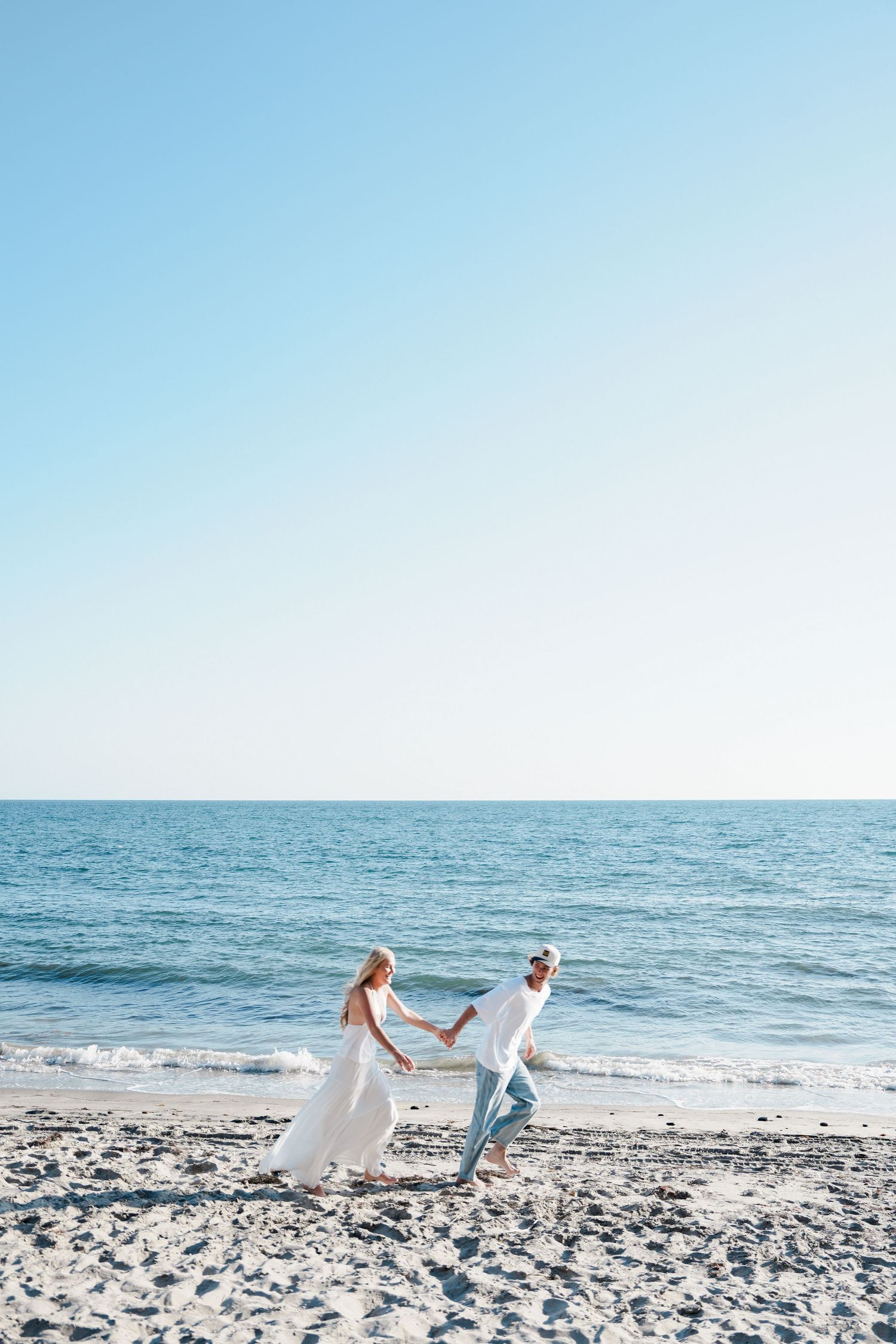 A bride and groom holding hands and dancing joyfully on a sunny beach with calm blue ocean waves behind them.