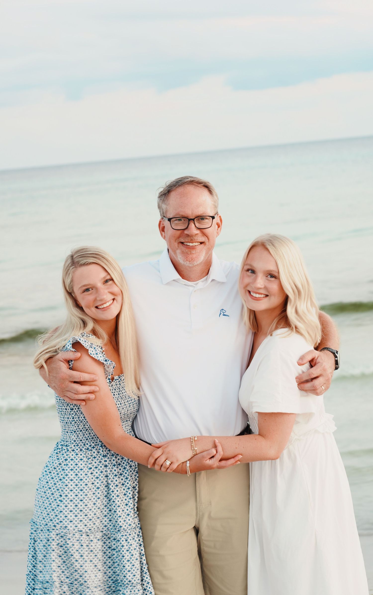 A family poses together in white and beige clothing at the beach with soft blue tones in the background.