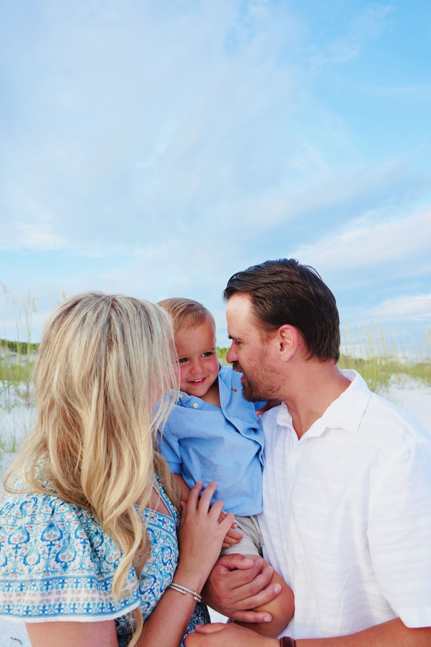 Family shares tender moments together at a sunny beach with white sand and blue sky background.