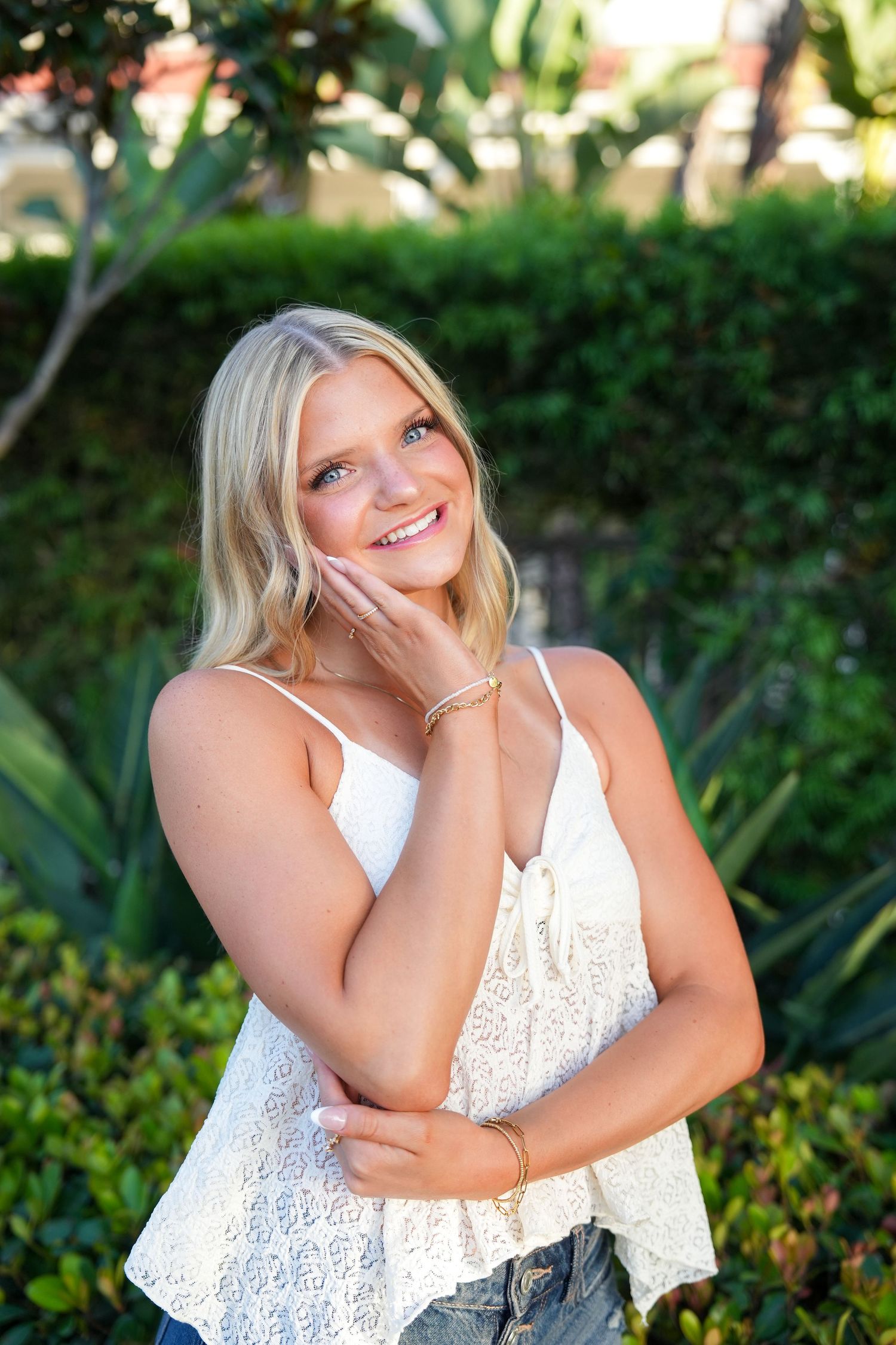 Smiling blonde woman in white lace top and denim shorts posing outdoors among lush green tropical plants.