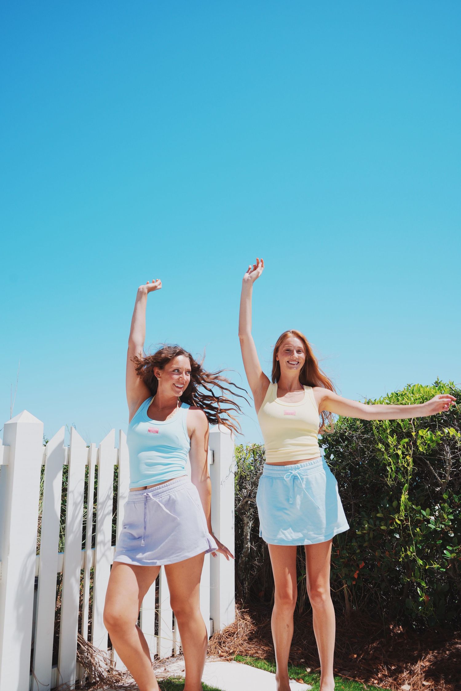 Two friends dancing and celebrating outdoors against bright blue sky and white fence.