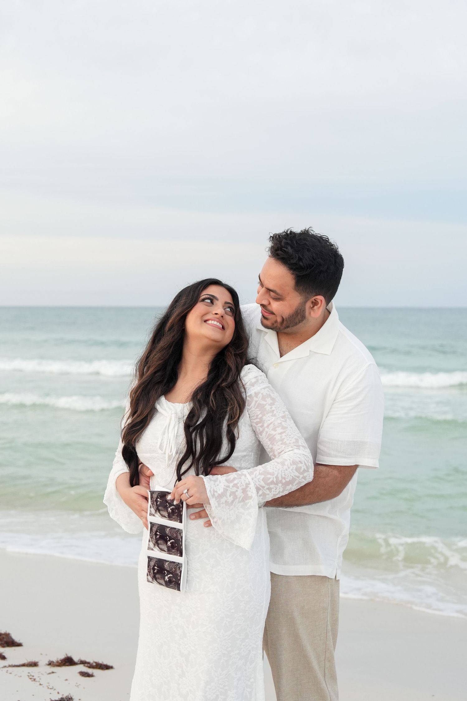 Couple in white outfits holding ultrasound photos on a beach, sharing a tender moment with ocean waves behind them.