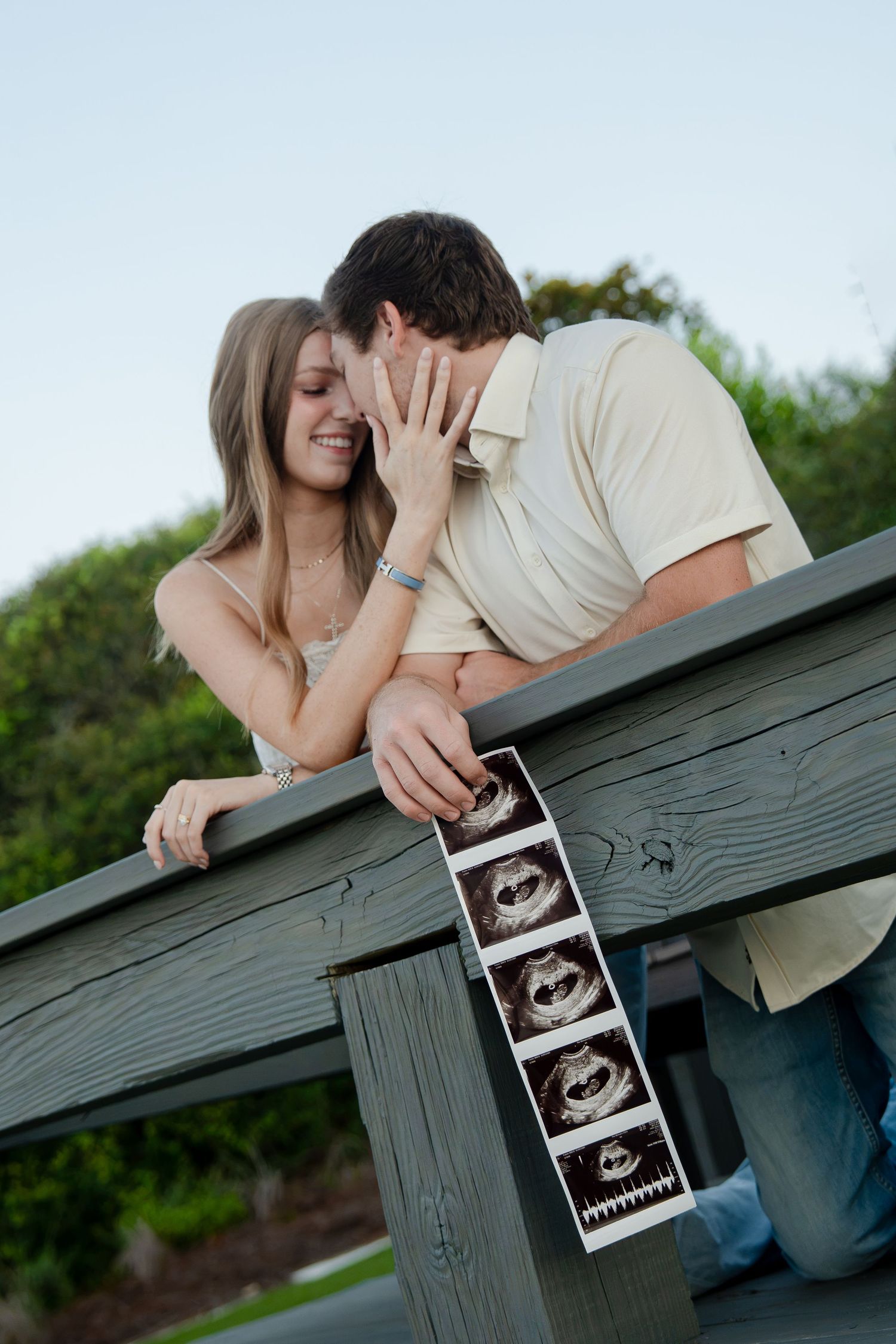 Happy couple sharing a kiss while holding ultrasound photos, announcing pregnancy outdoors on a wooden deck.