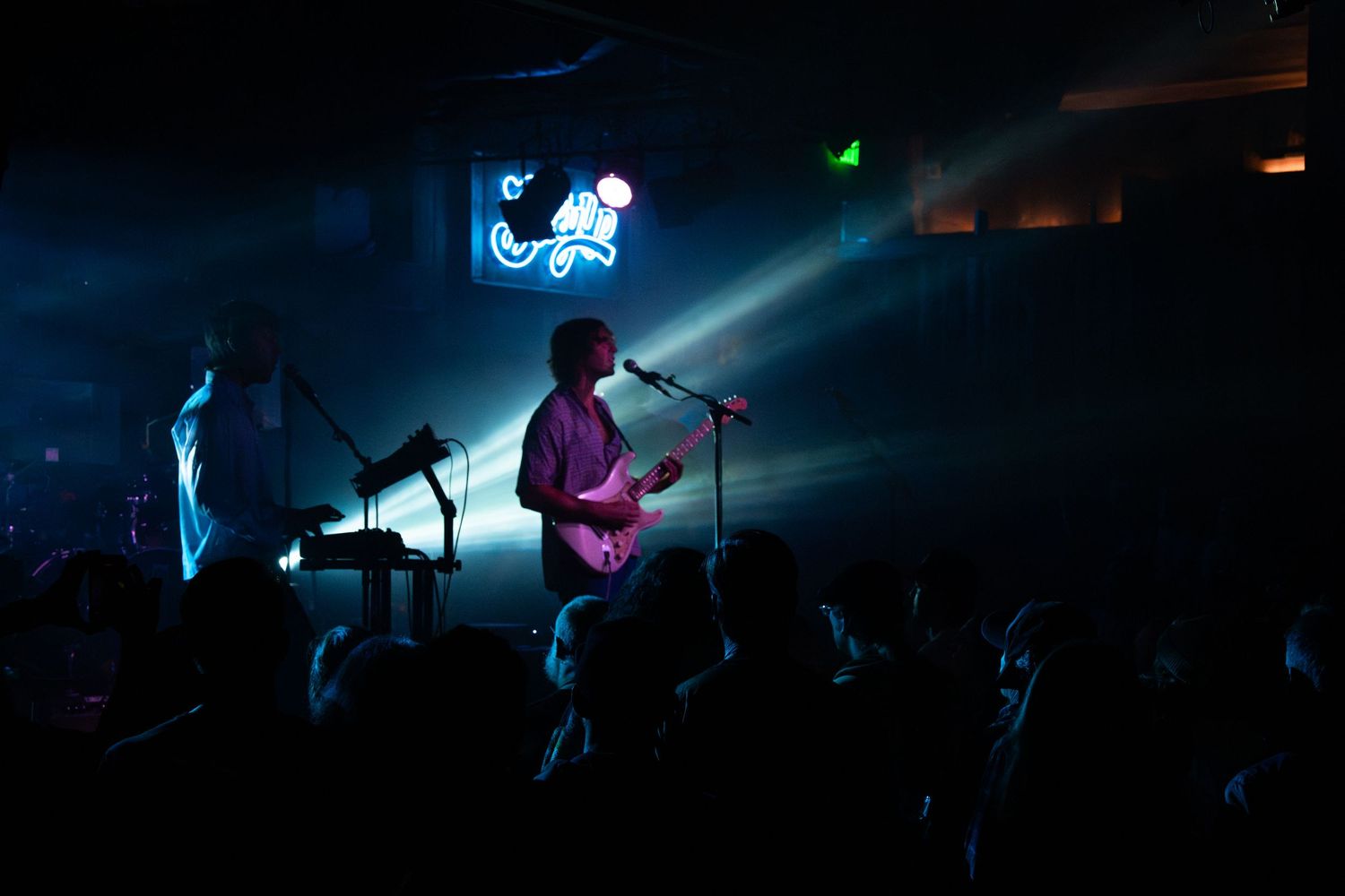 A rock band performs on a dark stage with blue neon lighting while silhouetted audience members watch from below.