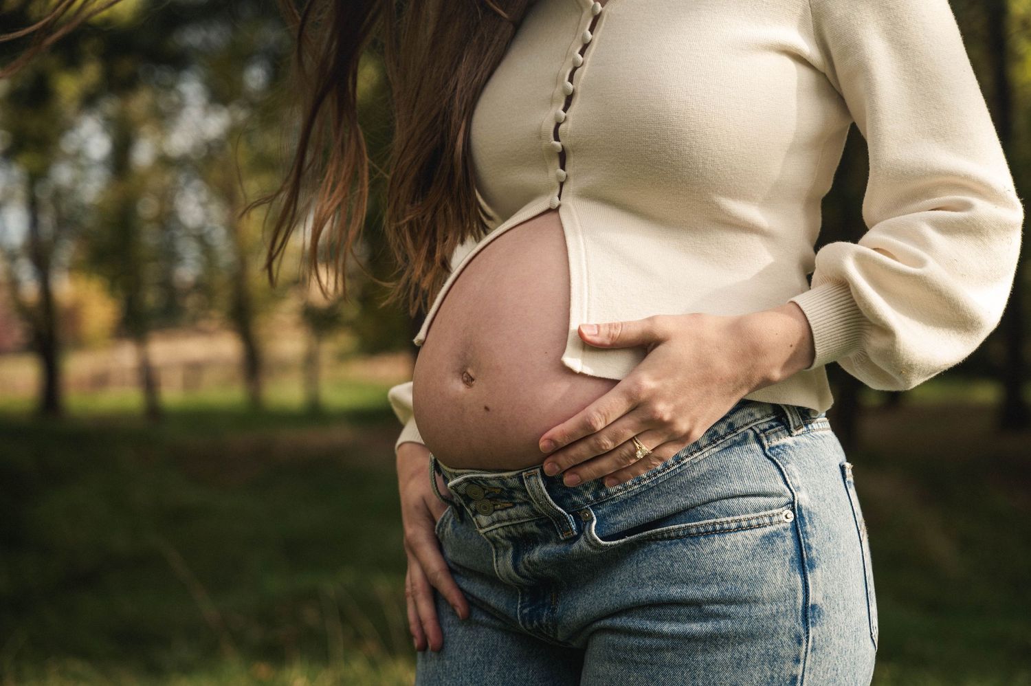 A pregnant woman in a cream sweater and jeans cradles her baby bump while standing outdoors in autumn.