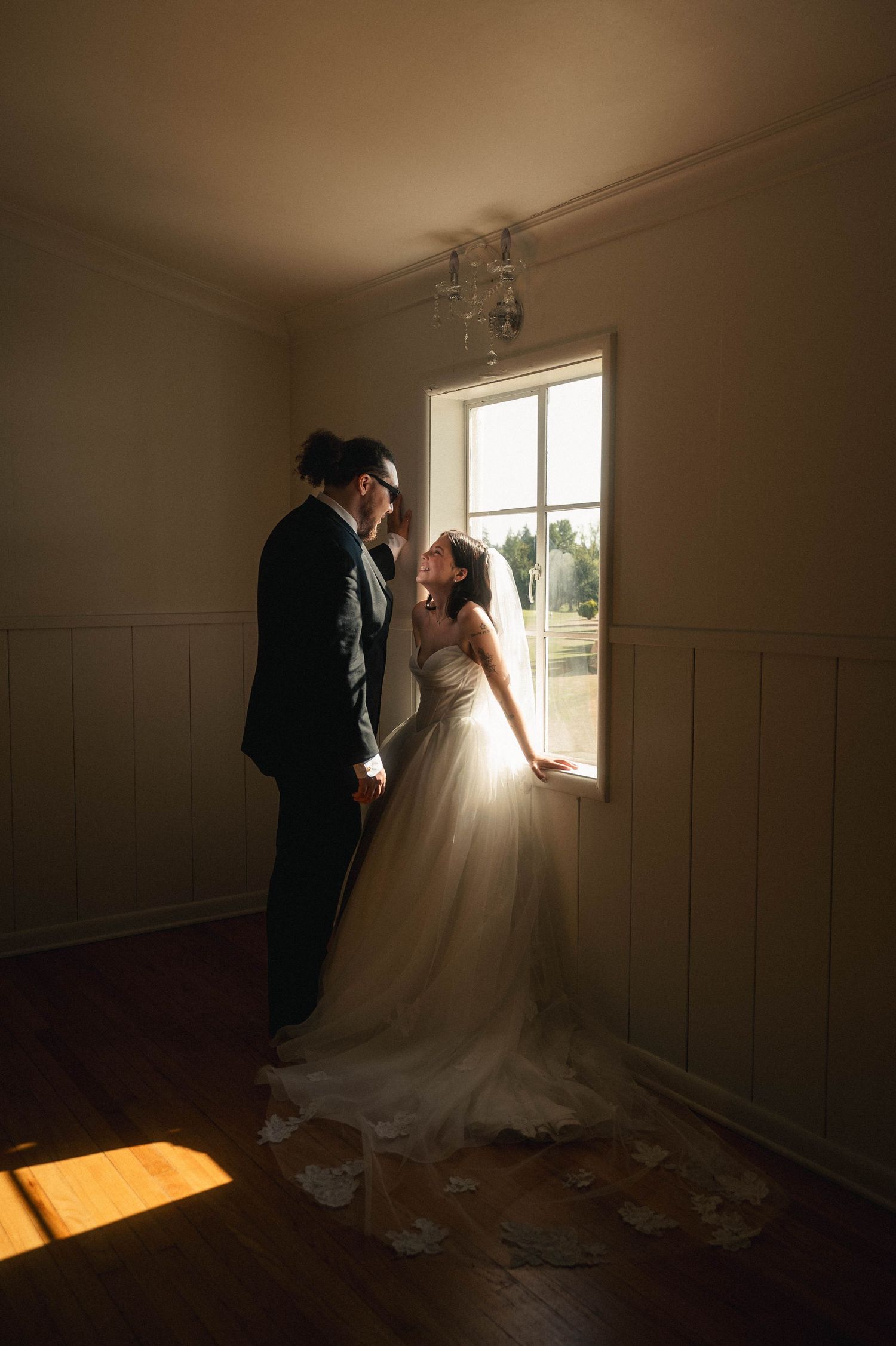 Bride and groom share a romantic moment by a sunlit window in a warmly lit room, bride in a flowing white gown.