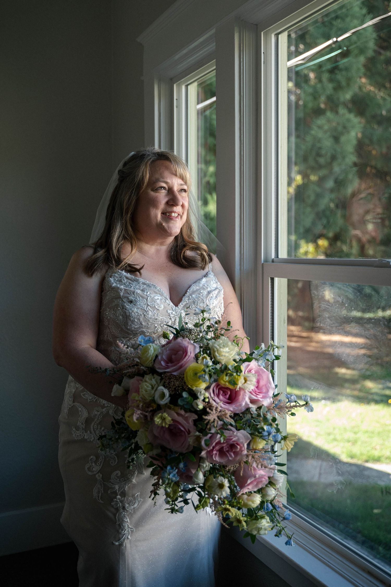 A bride in a lace wedding dress stands by a window holding a vibrant bouquet of pink and purple flowers.