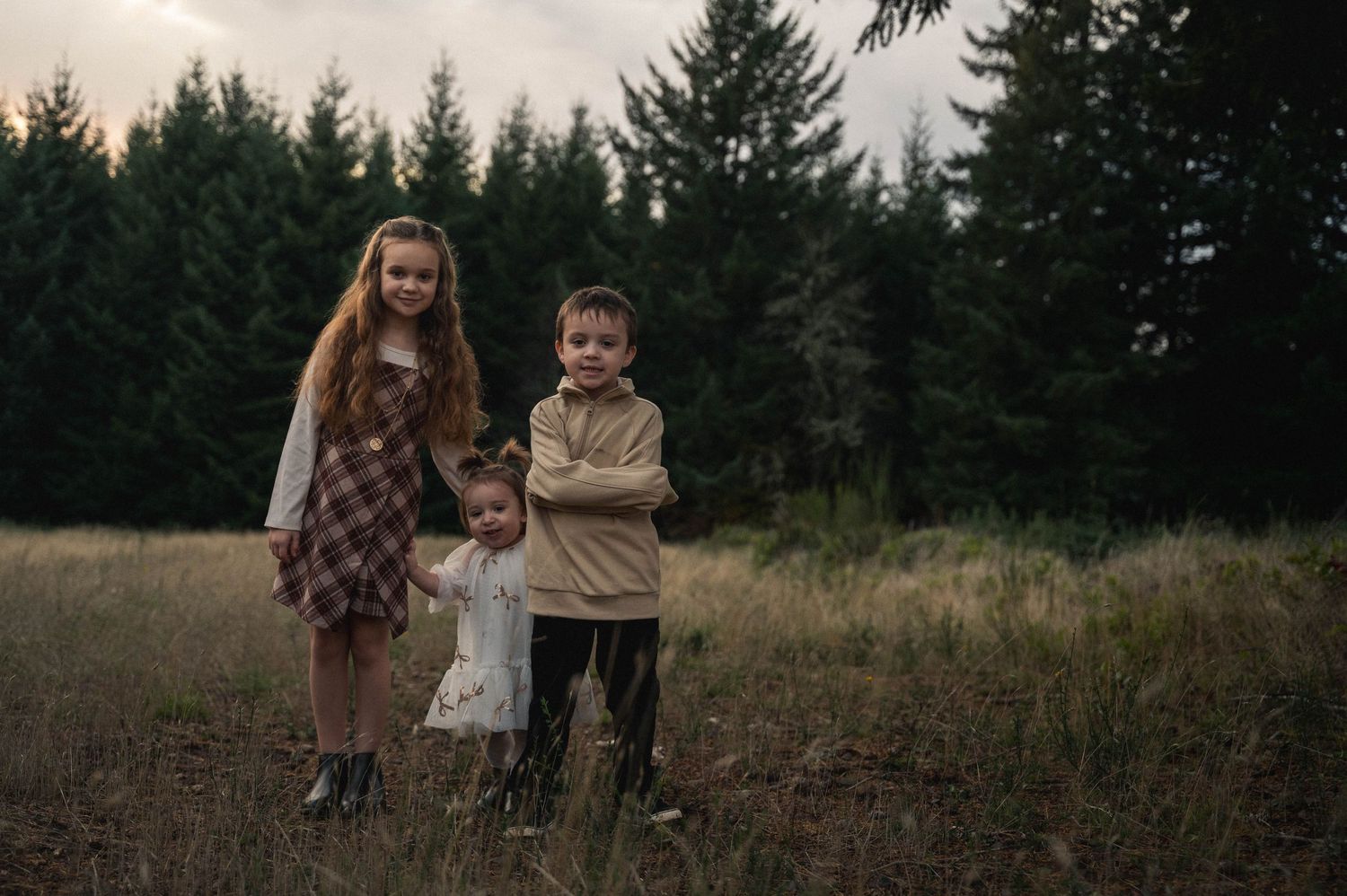A group stands together in a rustic outdoor field surrounded by evergreen trees at dusk.