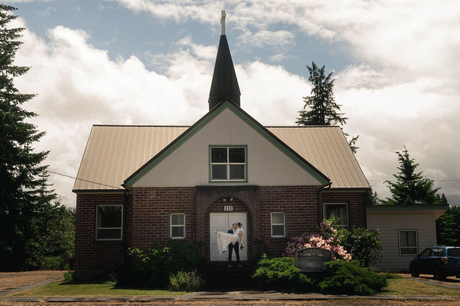 A couple stands at the entrance of a charming brick church with a metal roof and steeple, surrounded by lush trees.