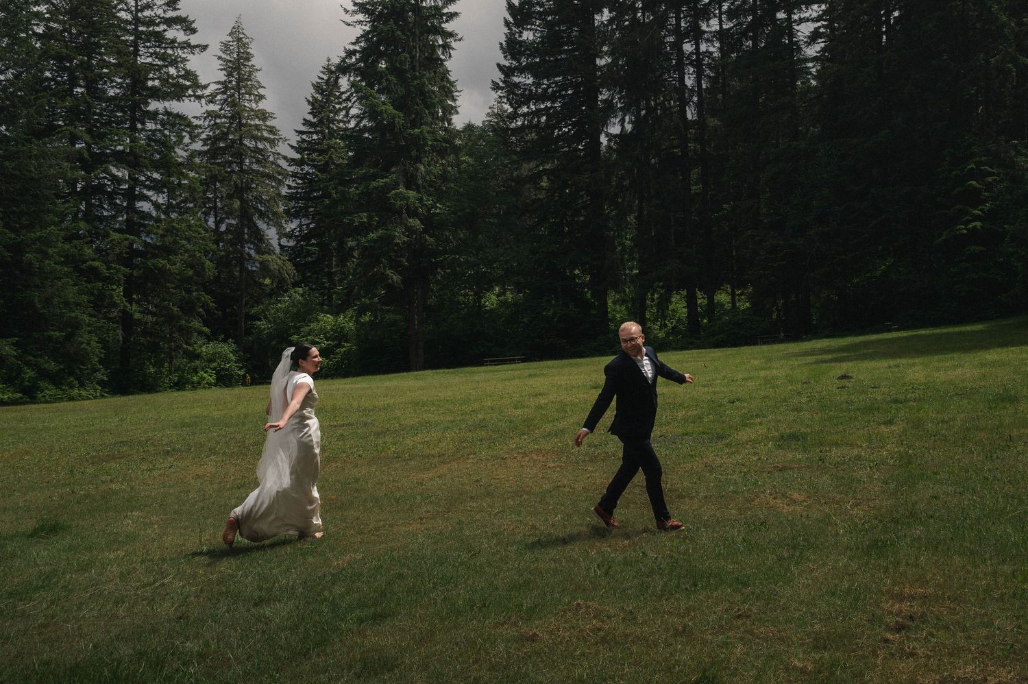 Bride and groom walking across lush green meadow surrounded by tall evergreen trees during outdoor forest wedding ceremony.