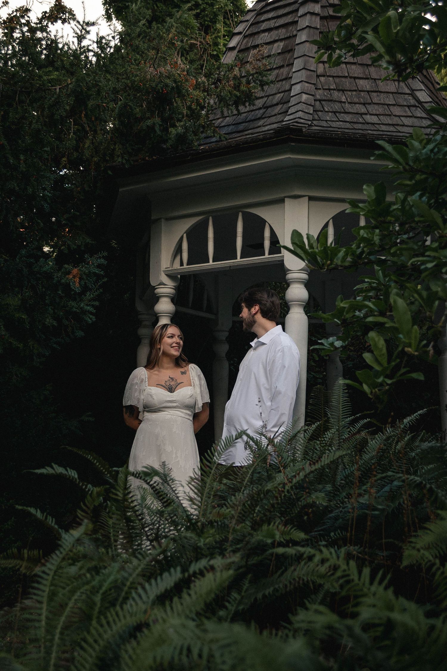 Man and woman, dressed in white, standing next to ferns under a pergola in Salem, Oregon.