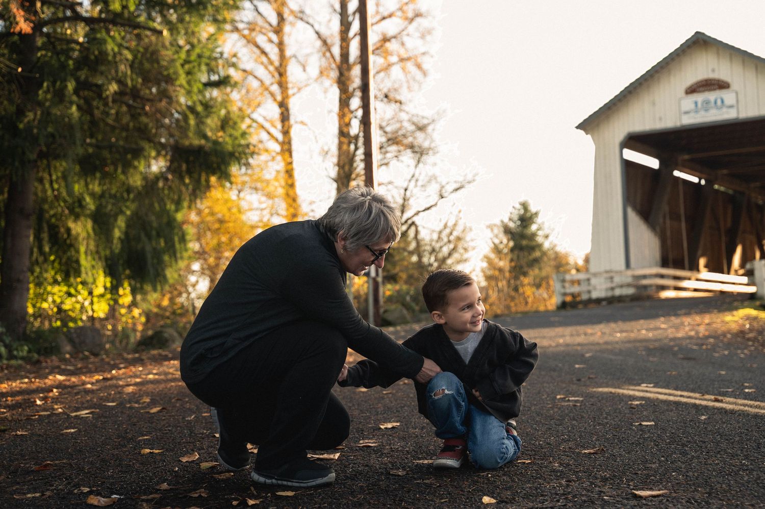 People sit on a gravel path during golden hour as autumn sunlight filters through trees in a peaceful outdoor setting.