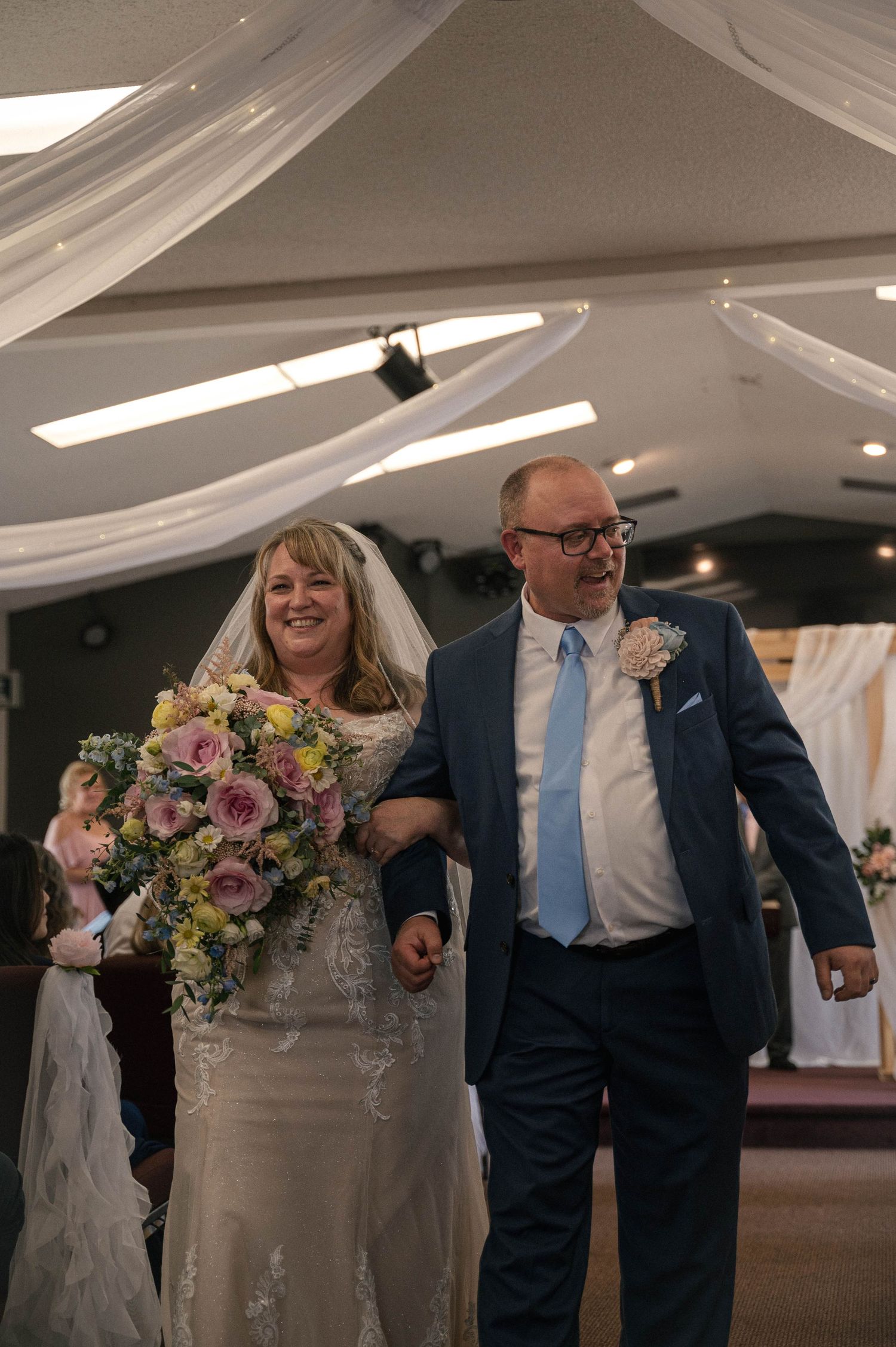 Wedding couple walks down aisle together in decorated venue with white draped ceiling and floral bouquet.