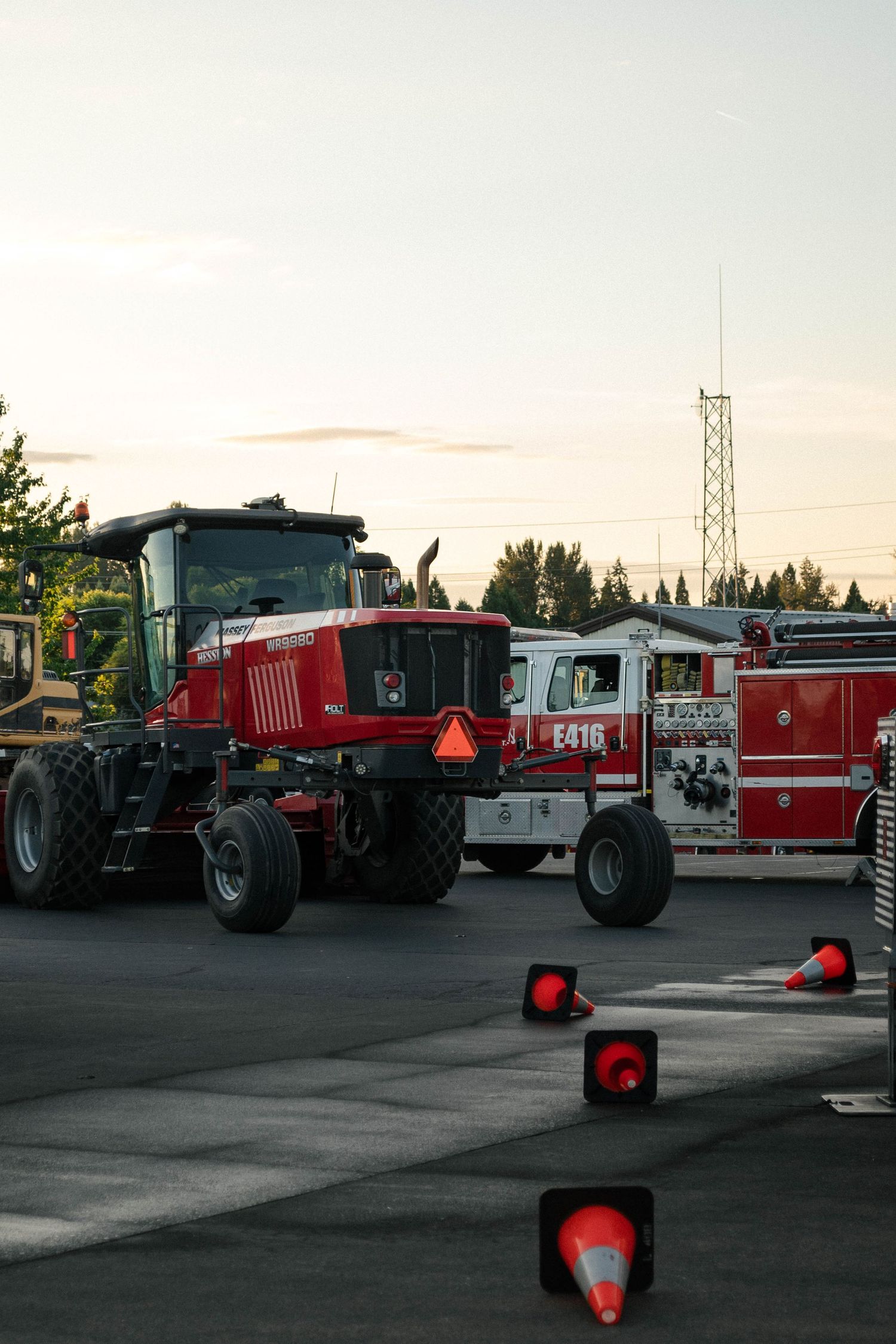 Red tractor and fire truck parked in lot at sunset with safety cones illuminated on the ground.