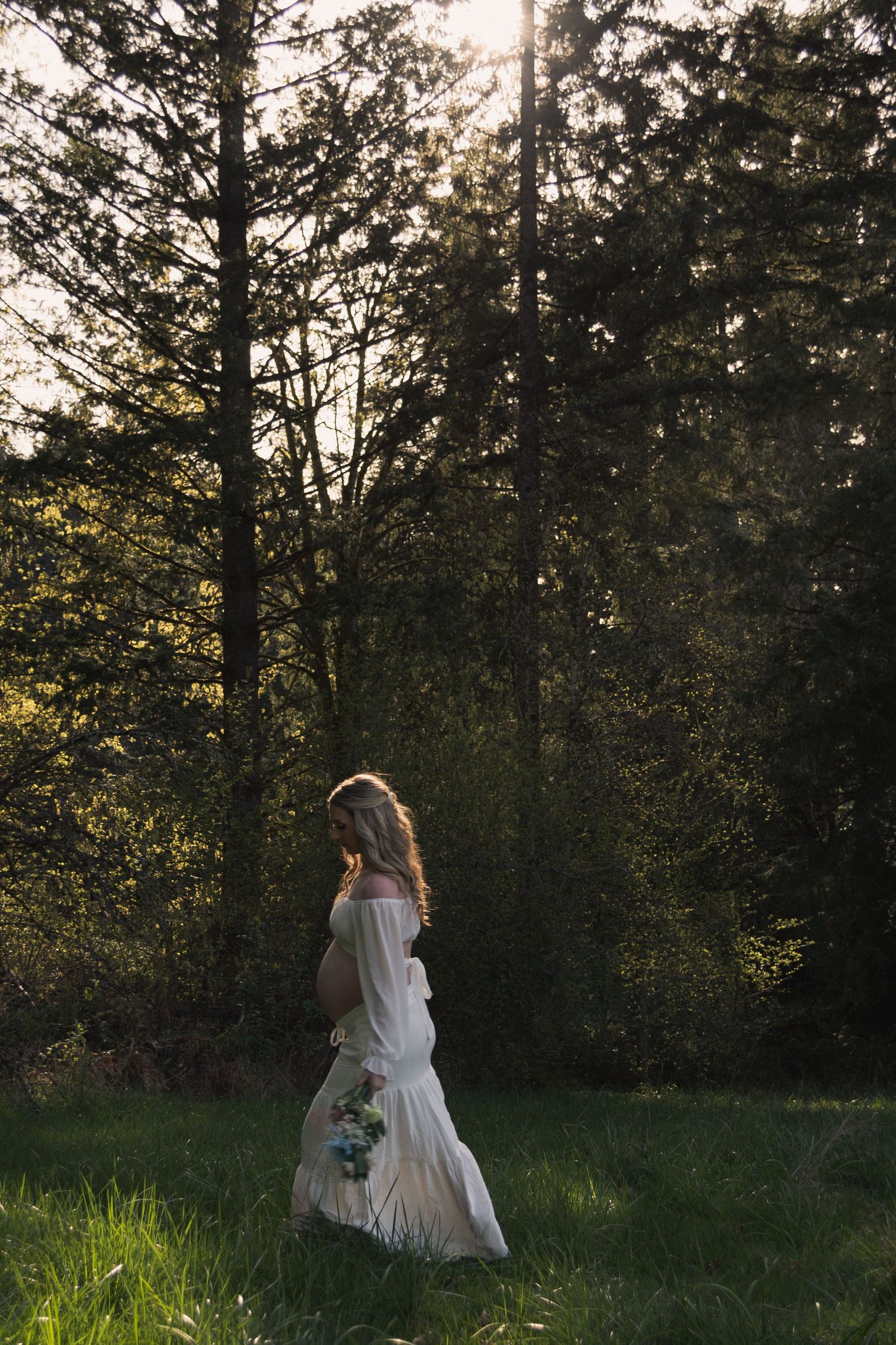 Person in flowing white dress walks through sunlit forest meadow.