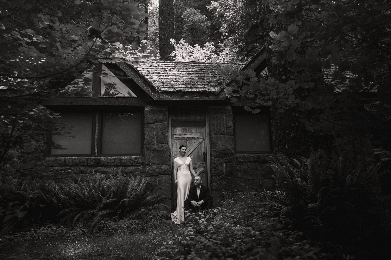 Black and white photo of a nude figure standing at the door of a rustic cottage surrounded by lush trees.