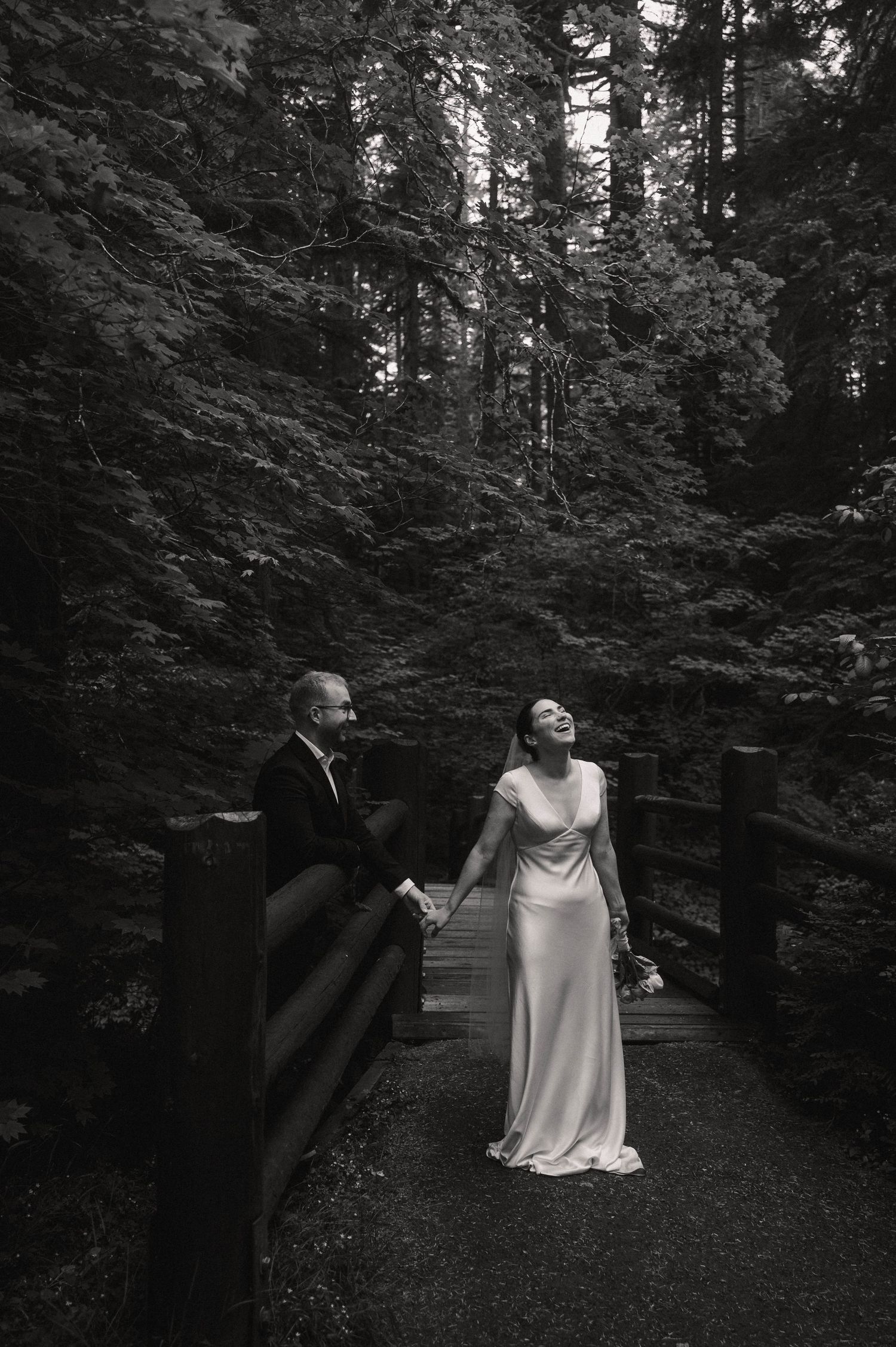 Black and white photo of bride laughing joyfully on a forest bridge with groom watching her during wedding day.