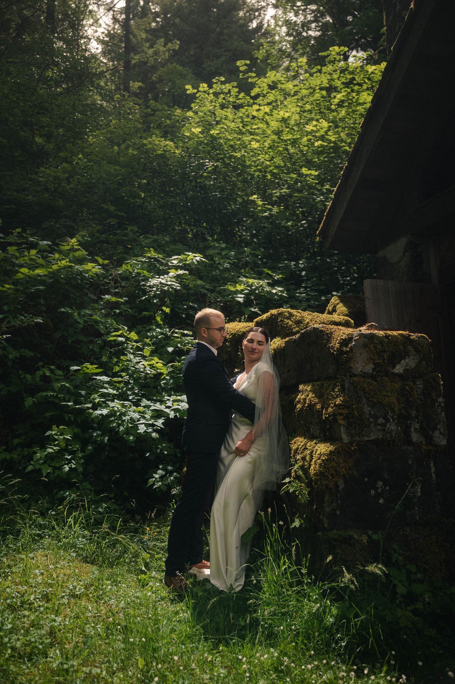 A bride and groom embrace near a moss-covered stone wall surrounded by lush green forest in an outdoor wedding portrait.