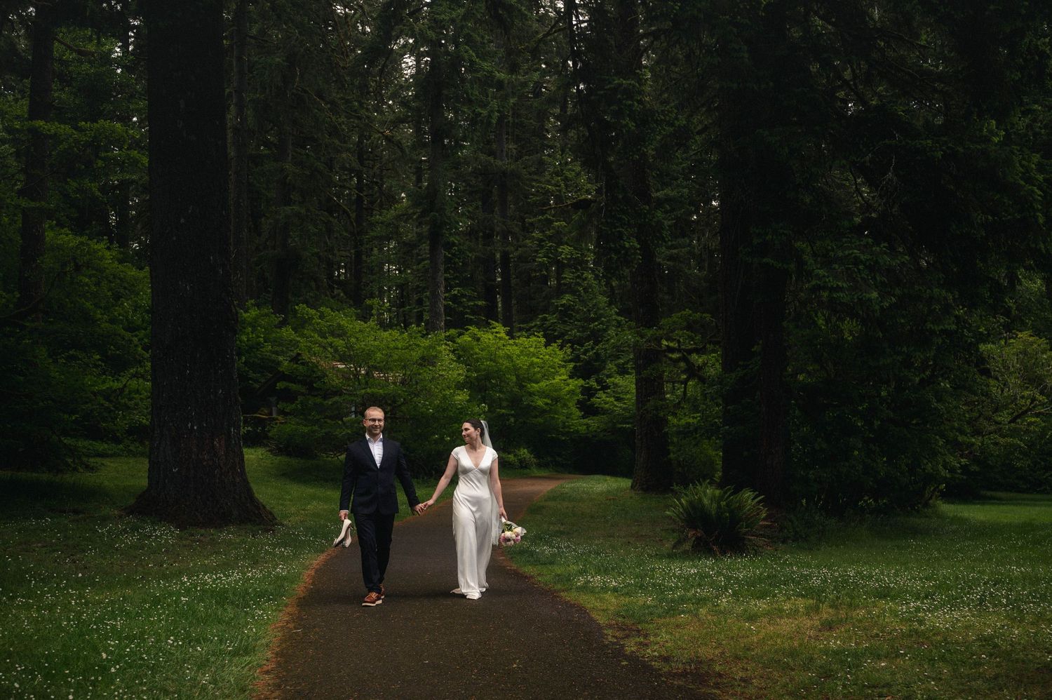 Bride and groom walk hand-in-hand along a forest path surrounded by tall evergreen trees on their wedding day.