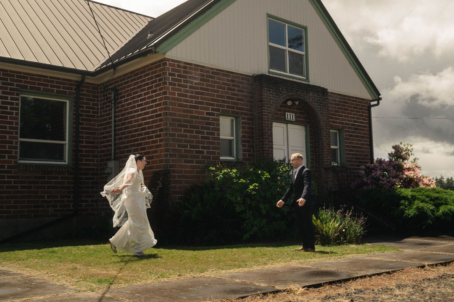 Bride in white gown and veil walks with groom in suit outside a brick church on a cloudy wedding day.
