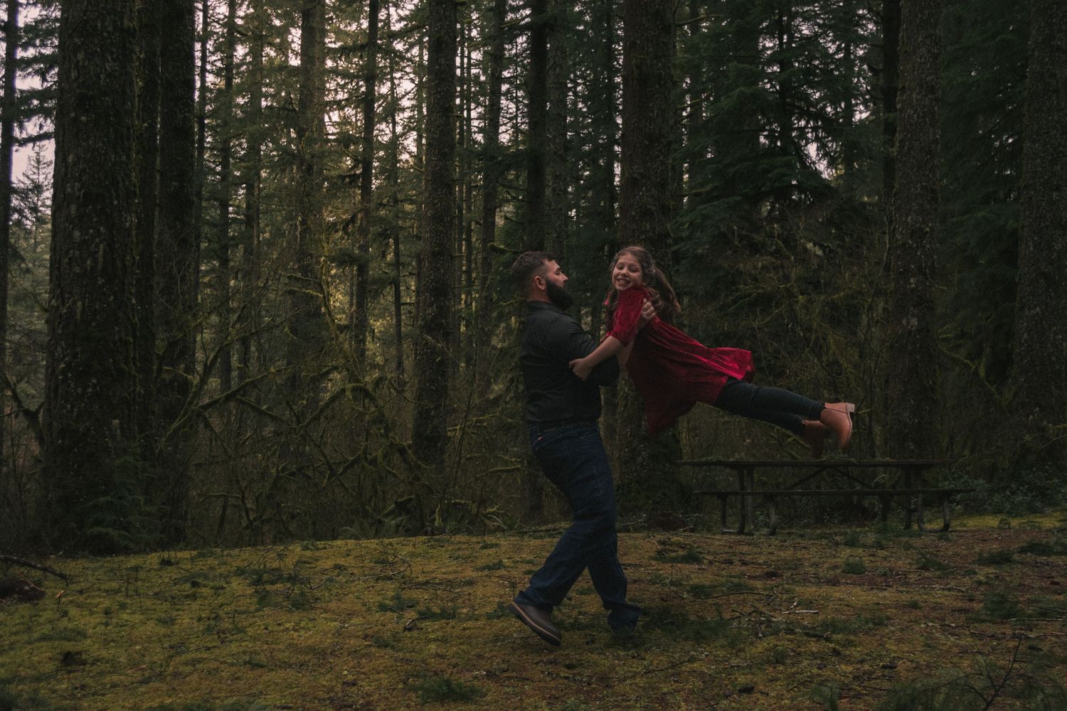 A couple shares a playful moment spinning in a moody forest setting.