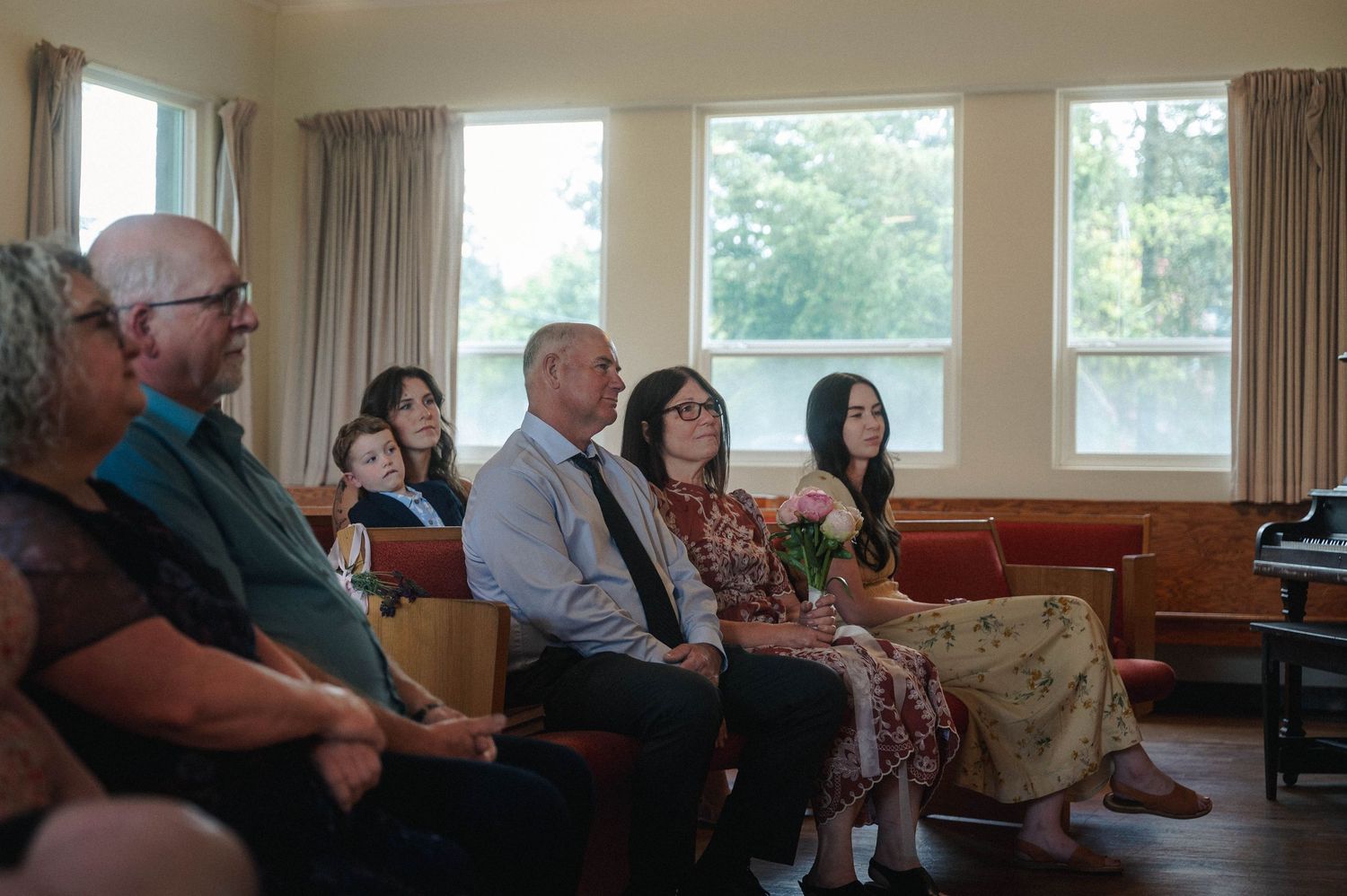 Guests seated in rows at an indoor ceremony, listening attentively in a bright room with large windows.