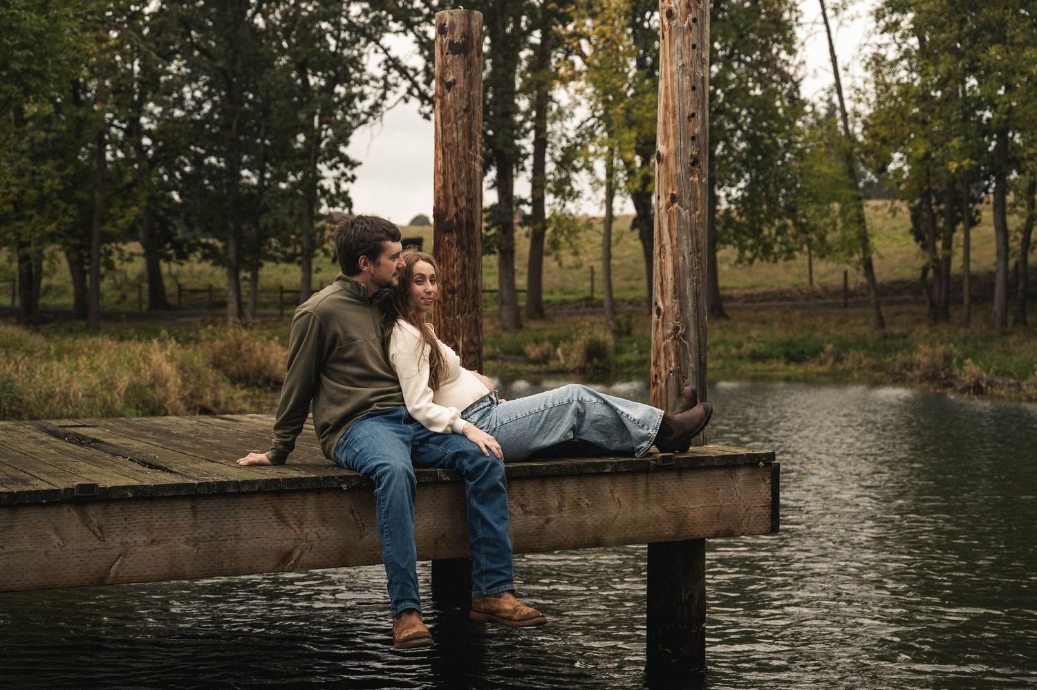 Pregnant woman and husband sitting close on a dock, feet hanging over the pond in Silverton, Oregon.
