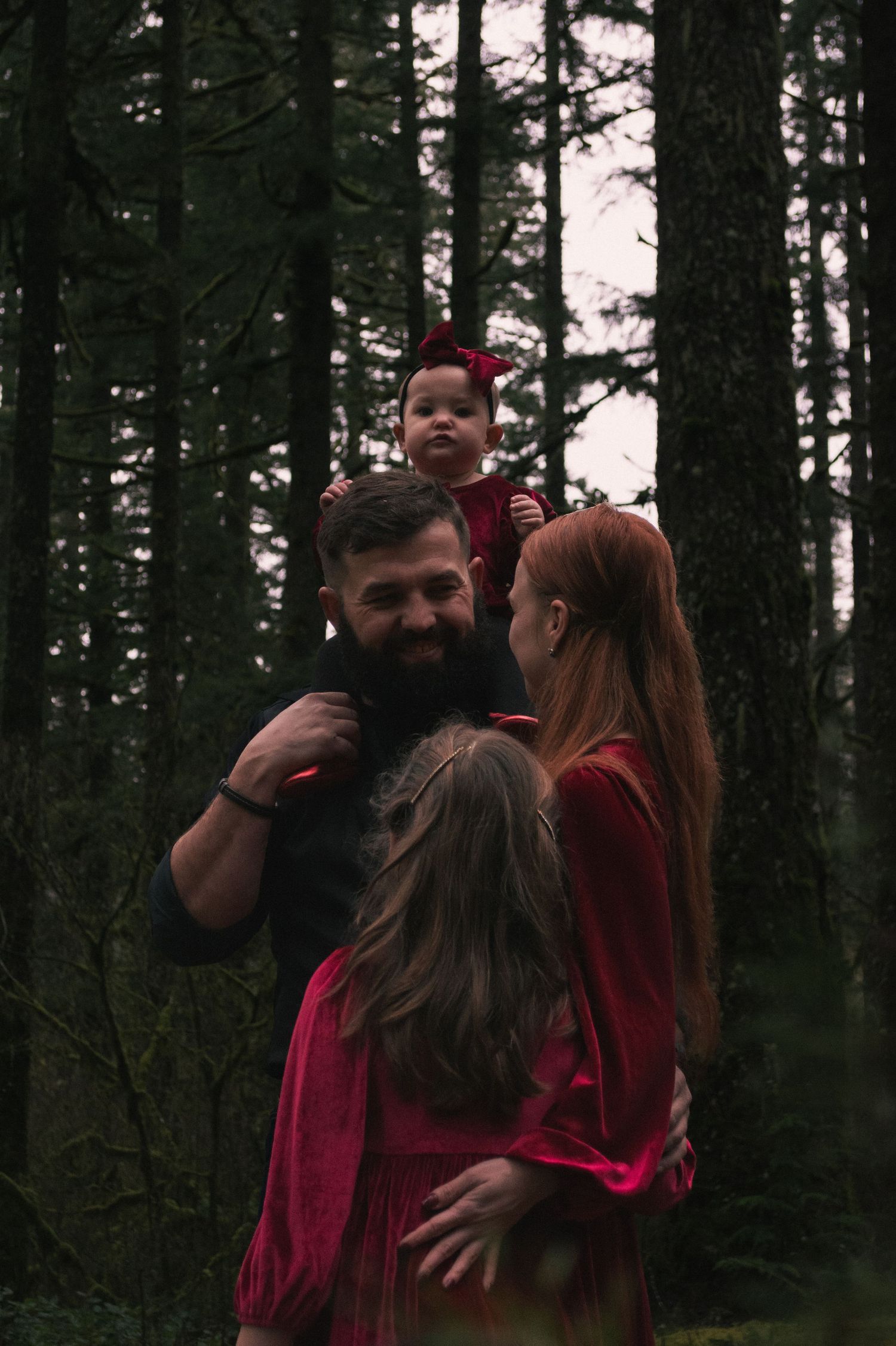 A family in red outfits shares an intimate moment together in a dark evergreen forest.