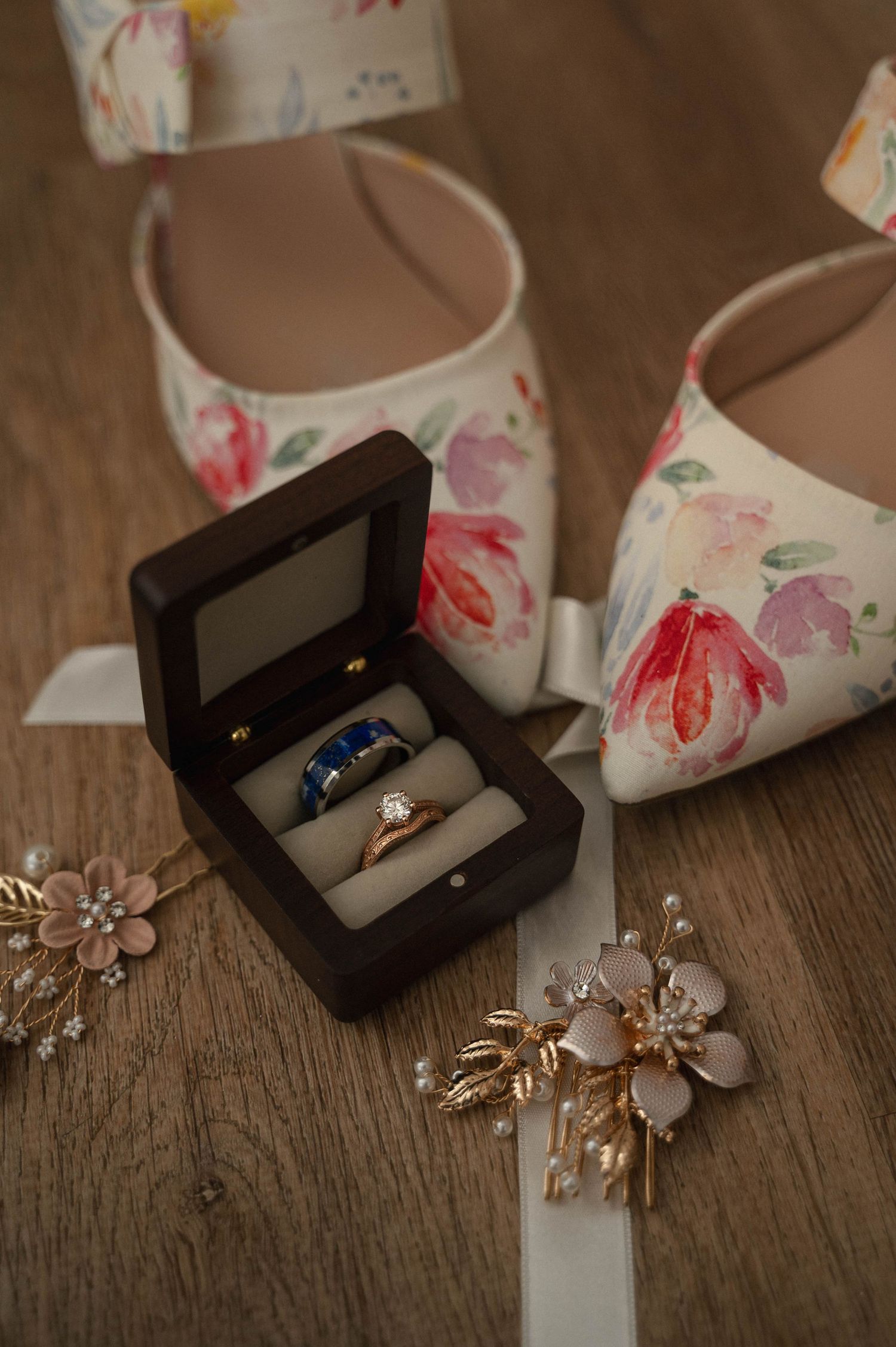 Floral white bridal shoes with delicate rose gold jewelry and ring boxes arranged on rustic wooden surface.