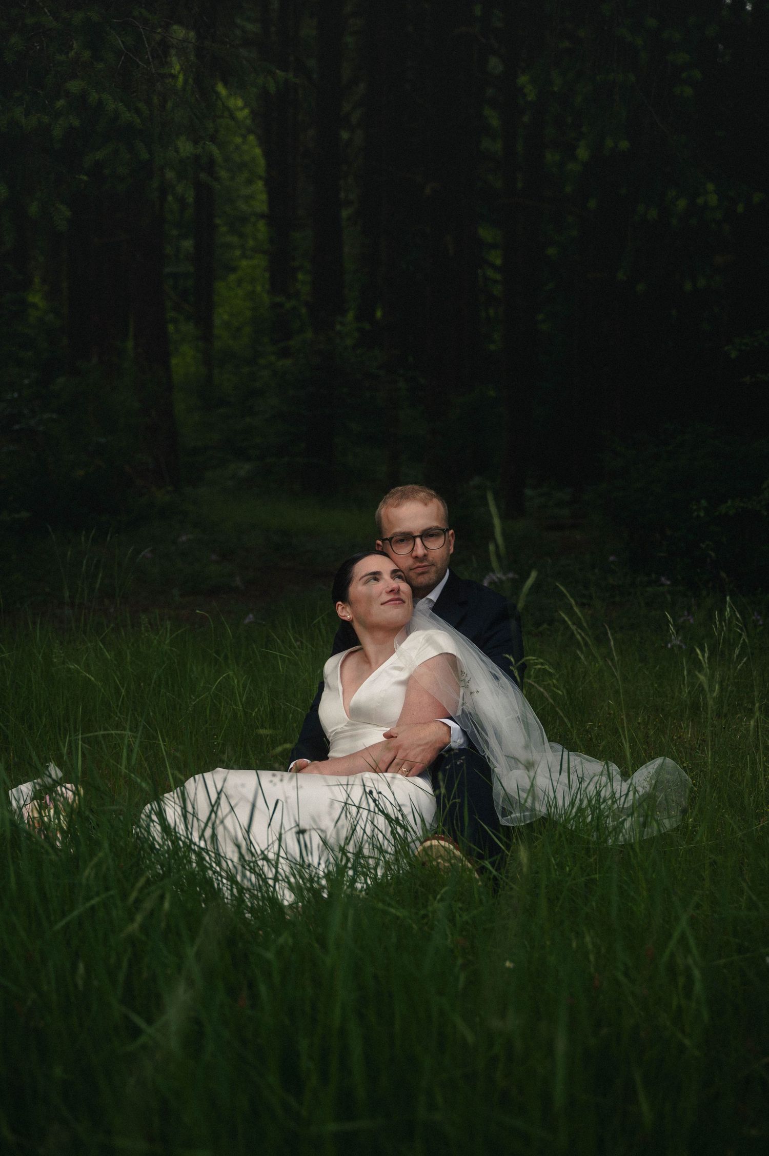 Bride and groom sit together in tall grass surrounded by a dark green forest in a romantic wedding portrait.