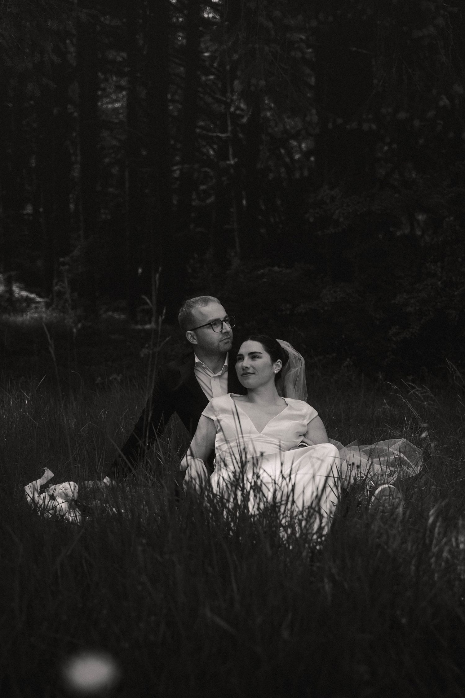 Black and white photo of a bride and groom sitting together in tall grass with a dark forest backdrop.