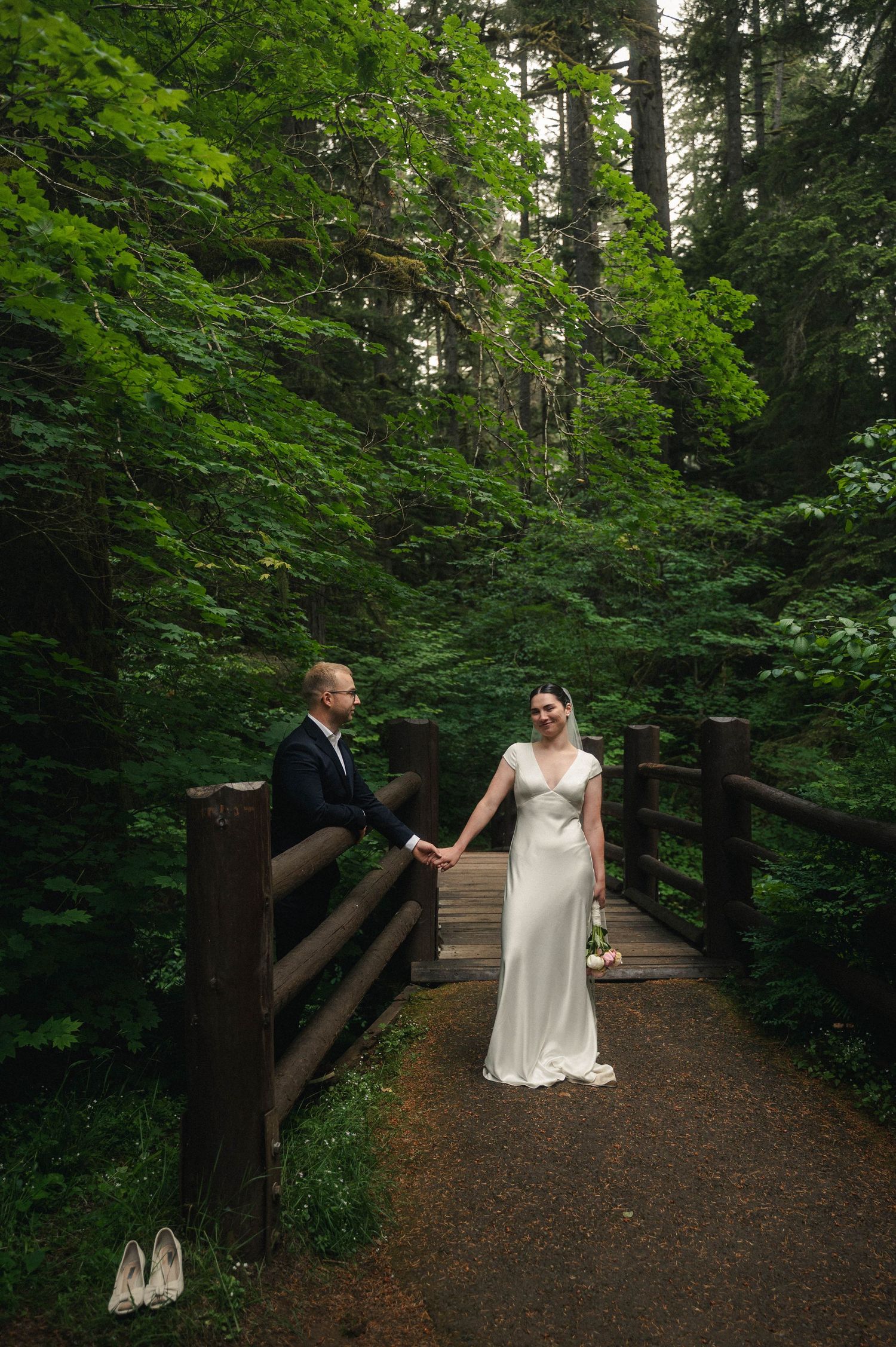 Bride and groom hold hands on a wooden bridge surrounded by lush green forest in a romantic outdoor wedding.