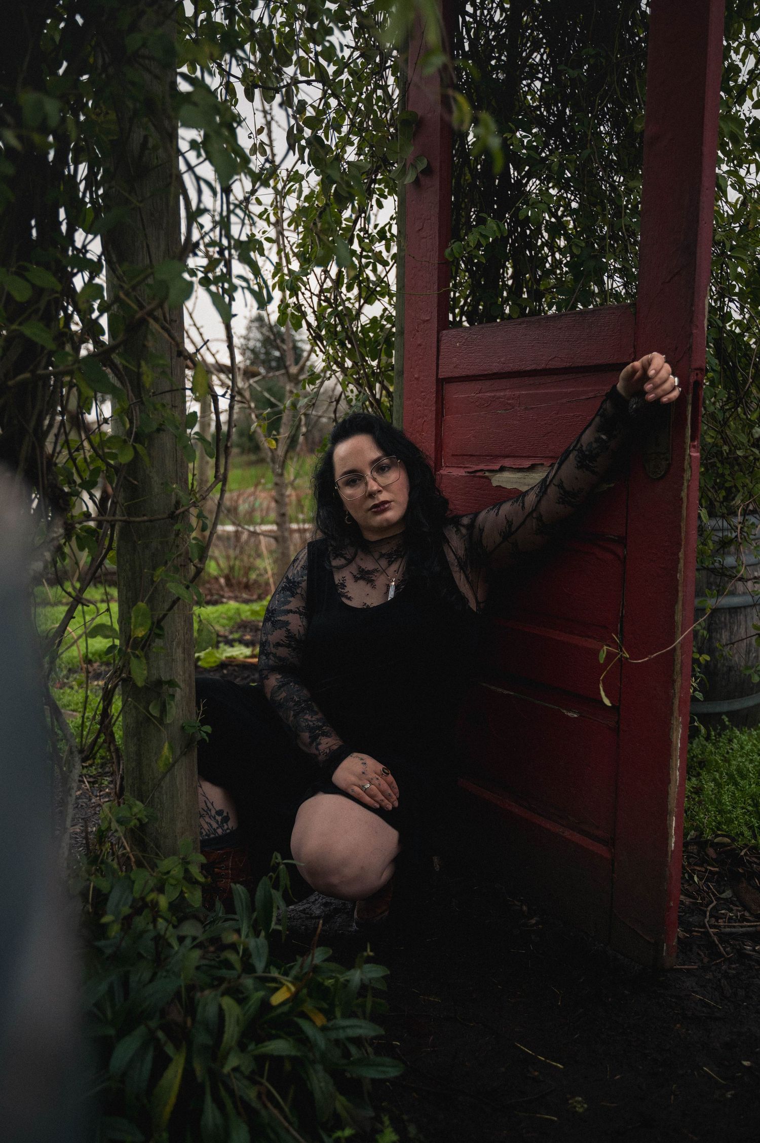 A person in black clothing poses by a red door surrounded by lush greenery in a moody outdoor setting.