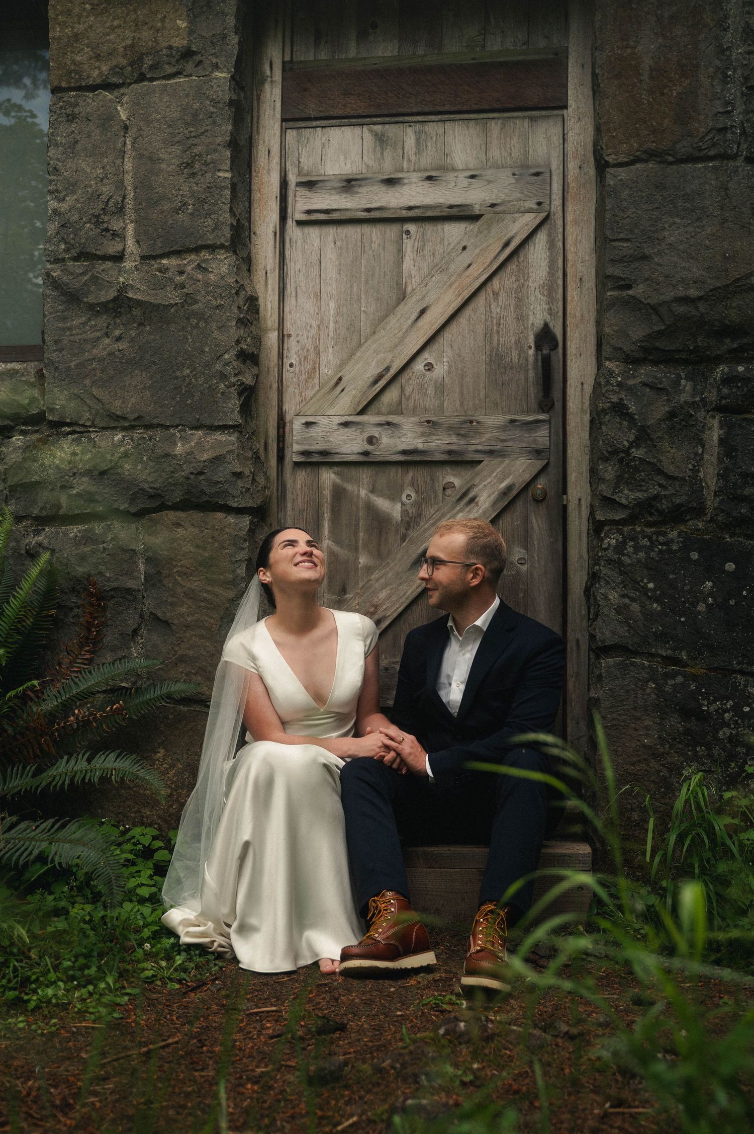 Bride and groom sit together laughing before a rustic wooden door on their wedding day.