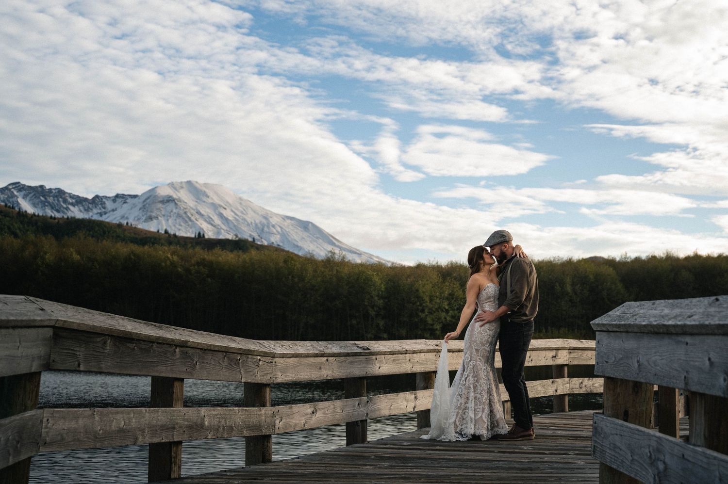 A couple shares an intimate moment on a rustic wooden boardwalk with majestic snow-capped mountains in the background.