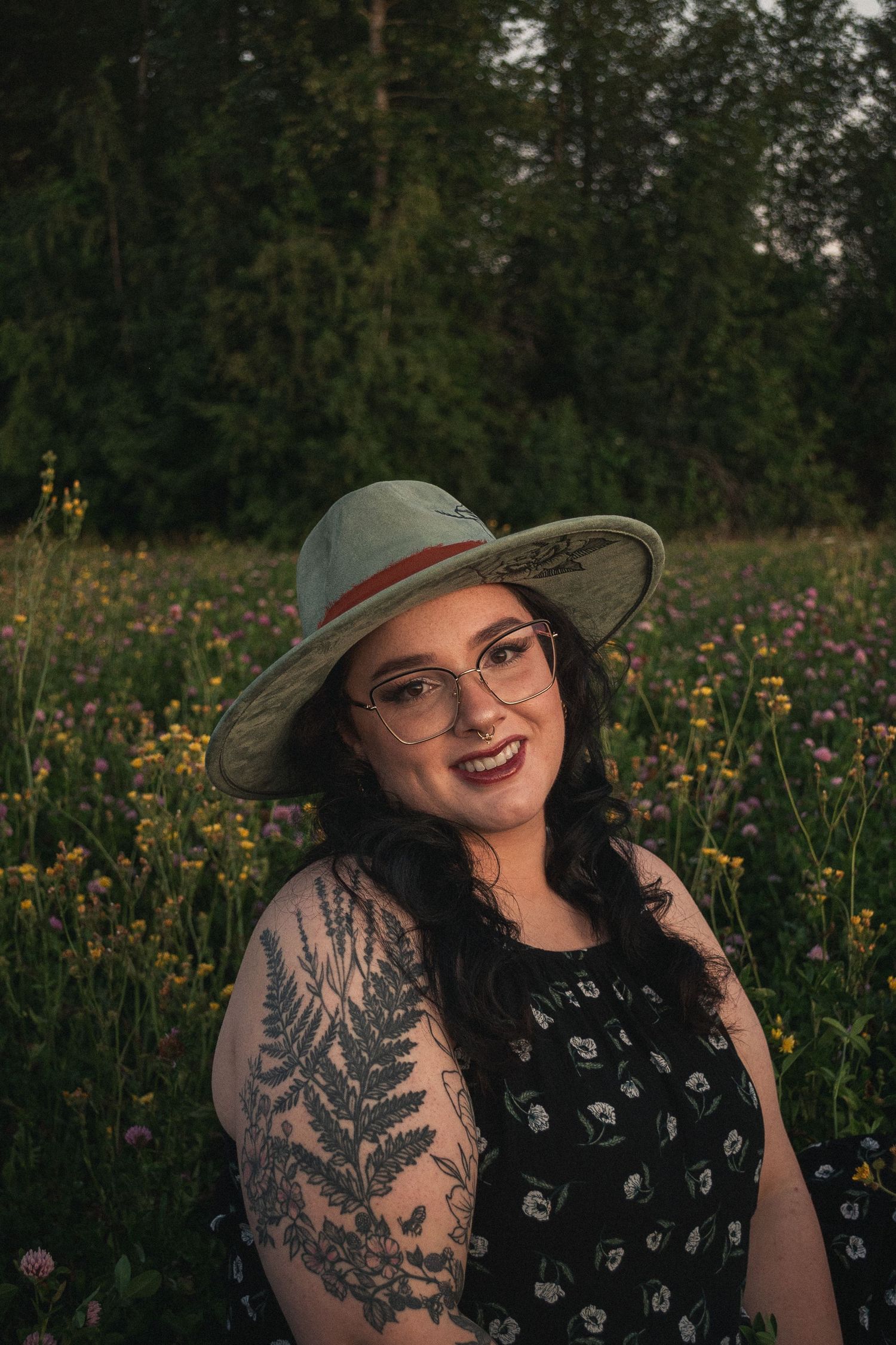 Woman with long dark hair, glasses, and round hat sitting posed in a clover field.