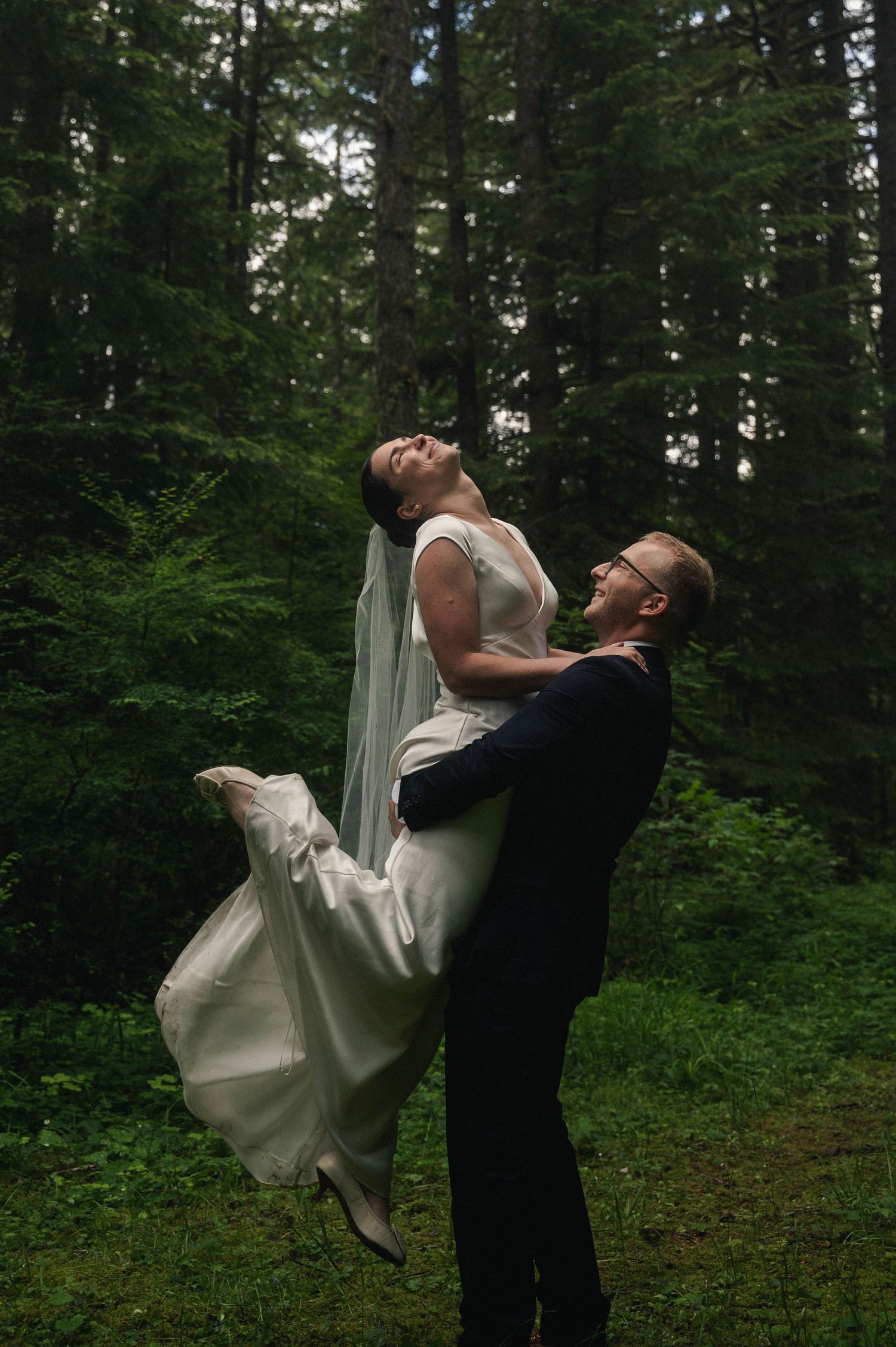 Groom lifts bride in white gown and veil, laughing joyfully in a lush green forest wedding photo.