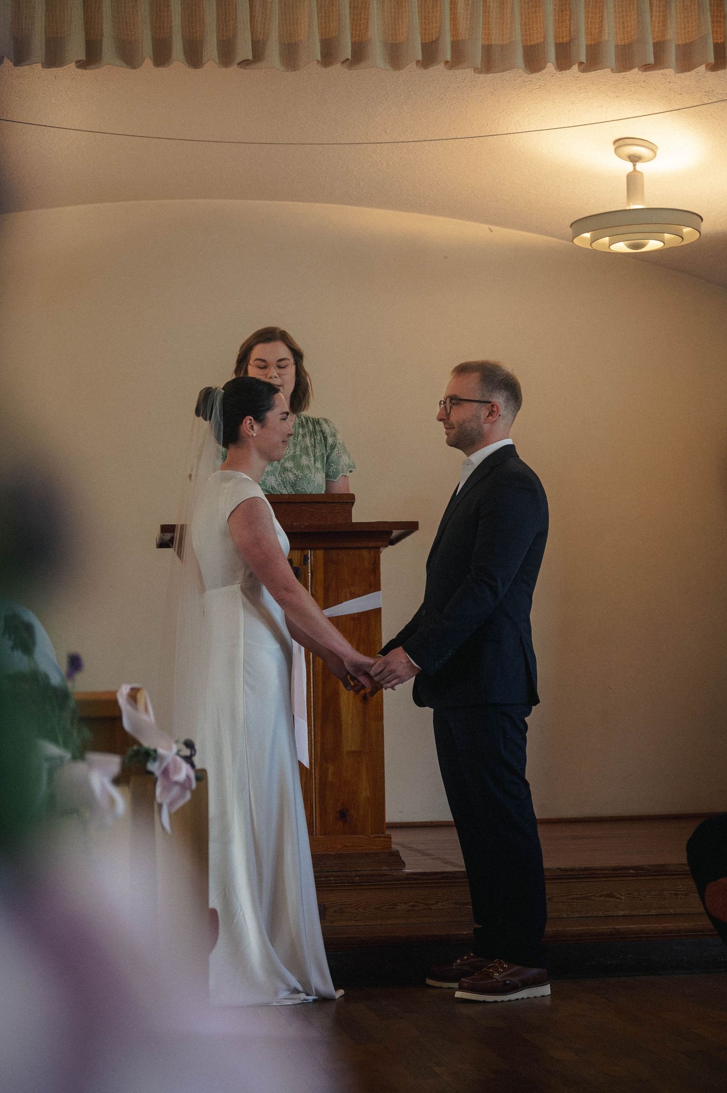 Bride and groom hold hands at altar during intimate wedding ceremony while officiant stands at wooden podium.