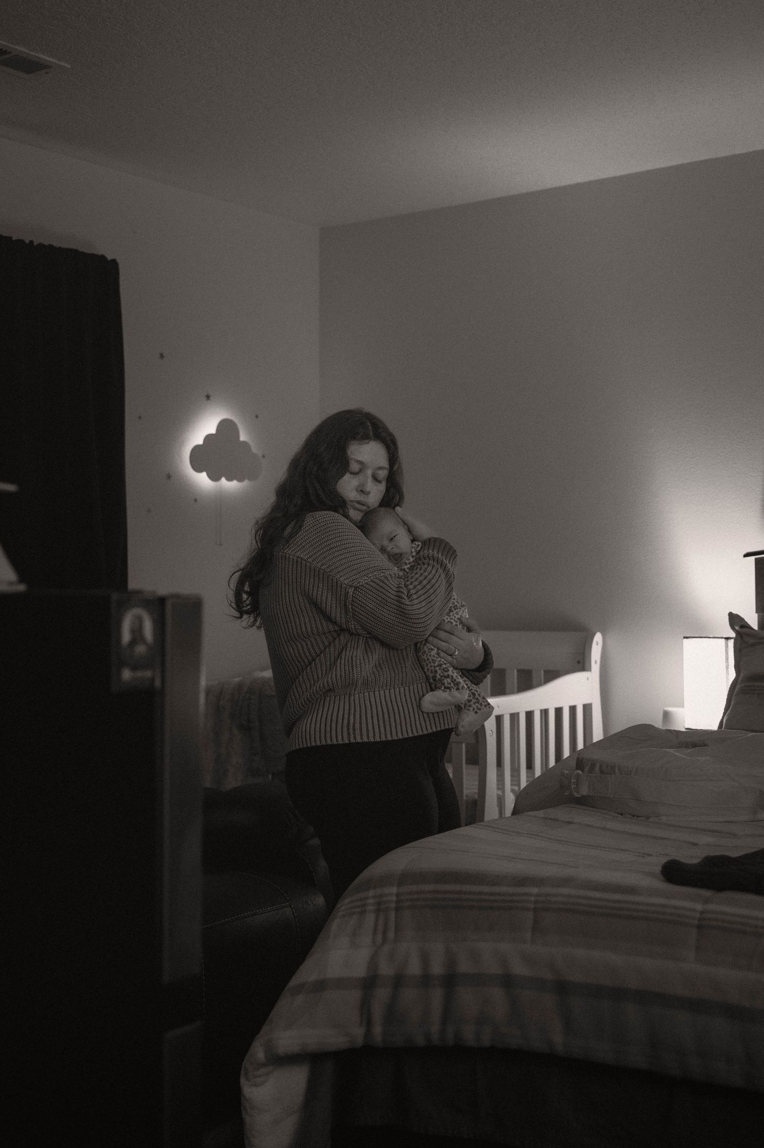 black and white image of mother, standing holding new born baby, rocking quietly next to a crib.