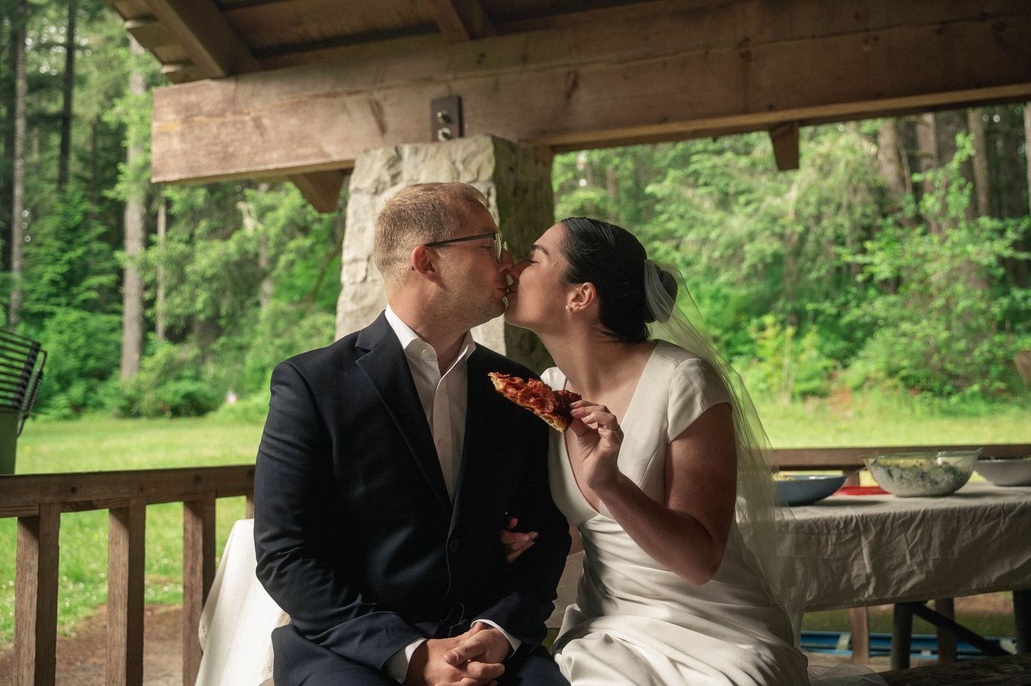 Newlywed couple sharing a kiss on a rustic wooden porch, bride in white dress holding a flower.