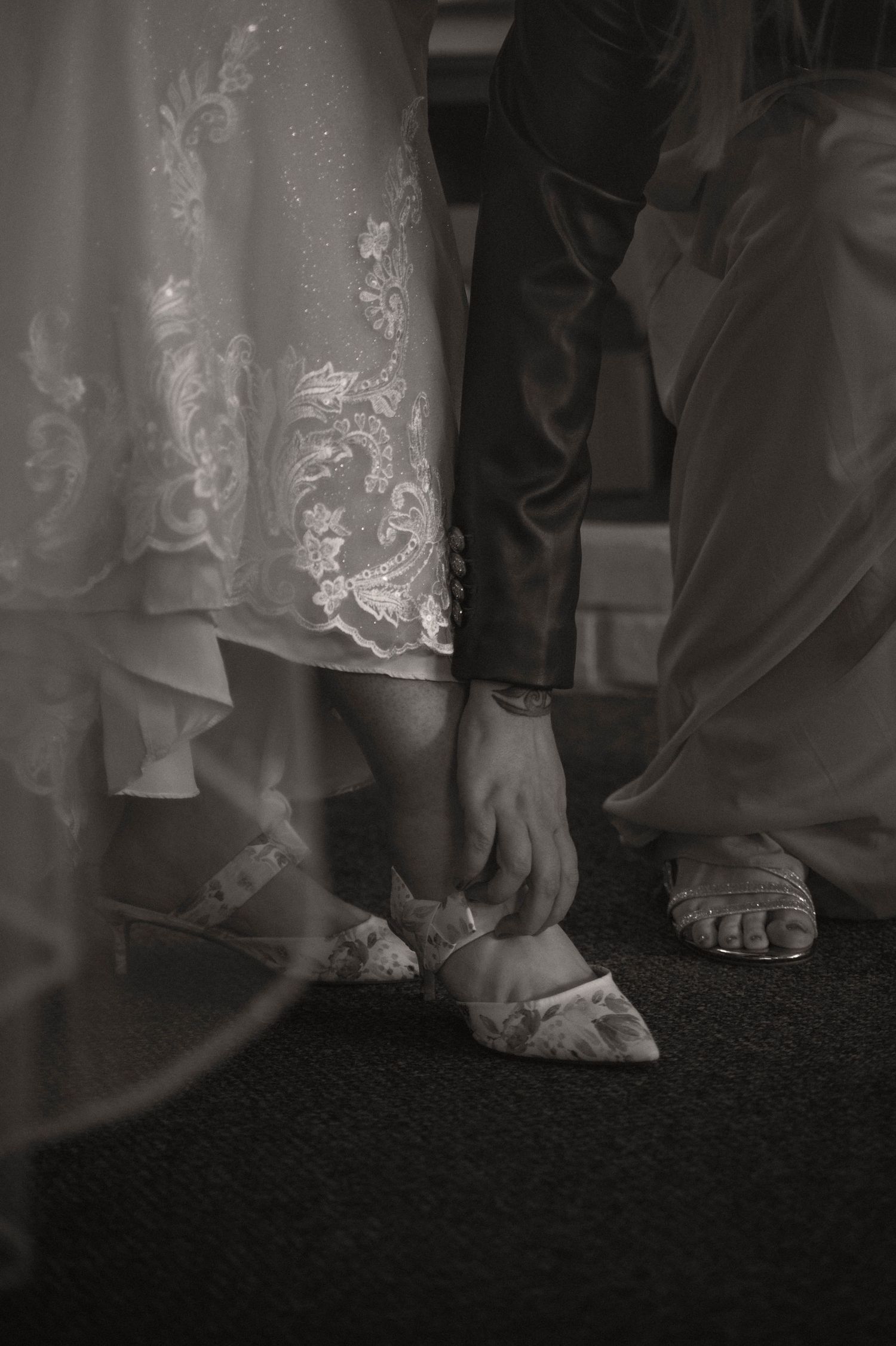 Close-up detail of white lace wedding dress hem and bare feet on dark carpet.