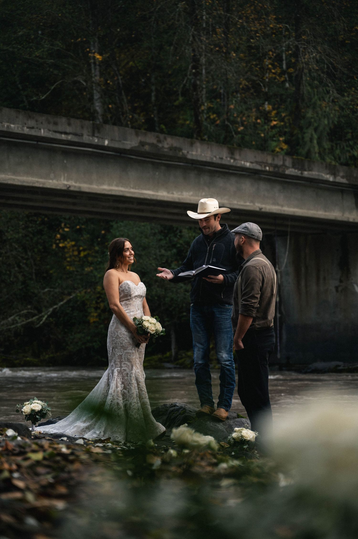A couple in wedding attire shares an intimate moment by a creek in a moody forest setting.