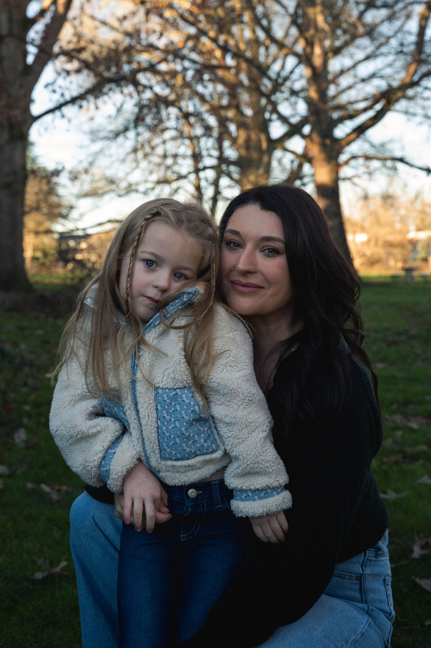 A parent and child wearing matching denim and cozy sweaters sit together outdoors in autumn sunlight.
