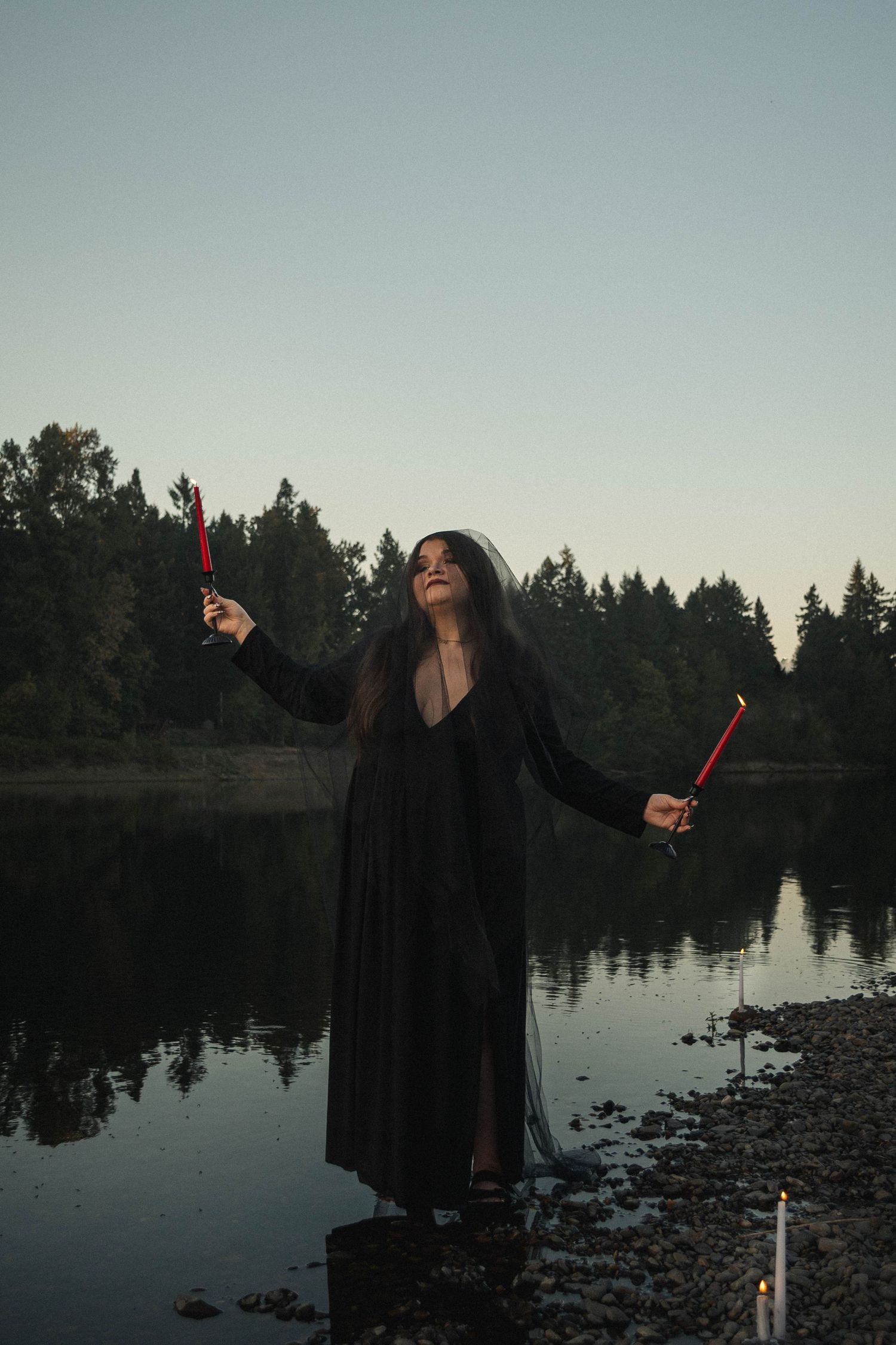 Figure in flowing black dress performs dramatic poses by a serene lake at dusk.