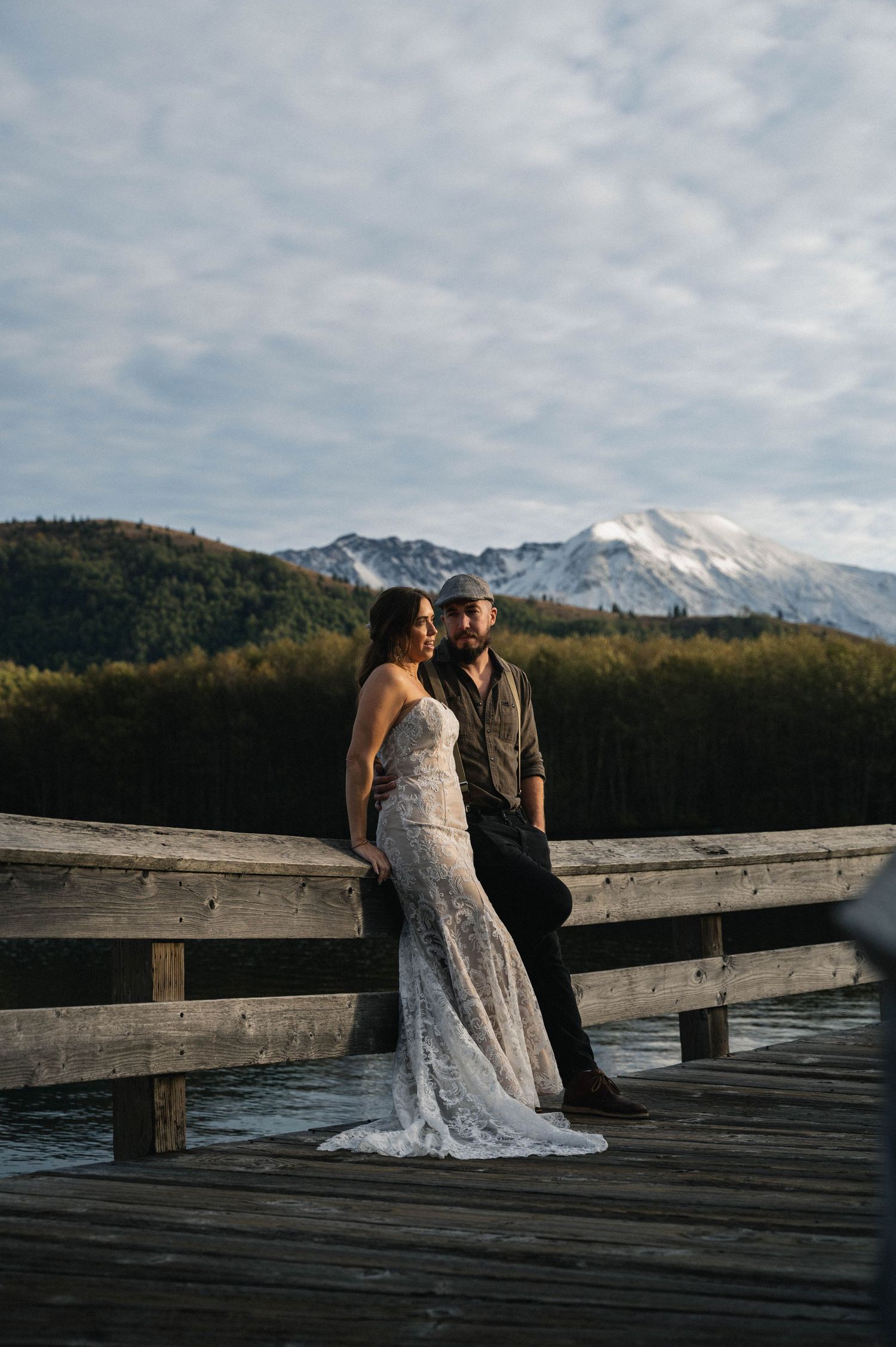 A couple stands together on a wooden dock with snow-capped mountains and cloudy skies in the background.