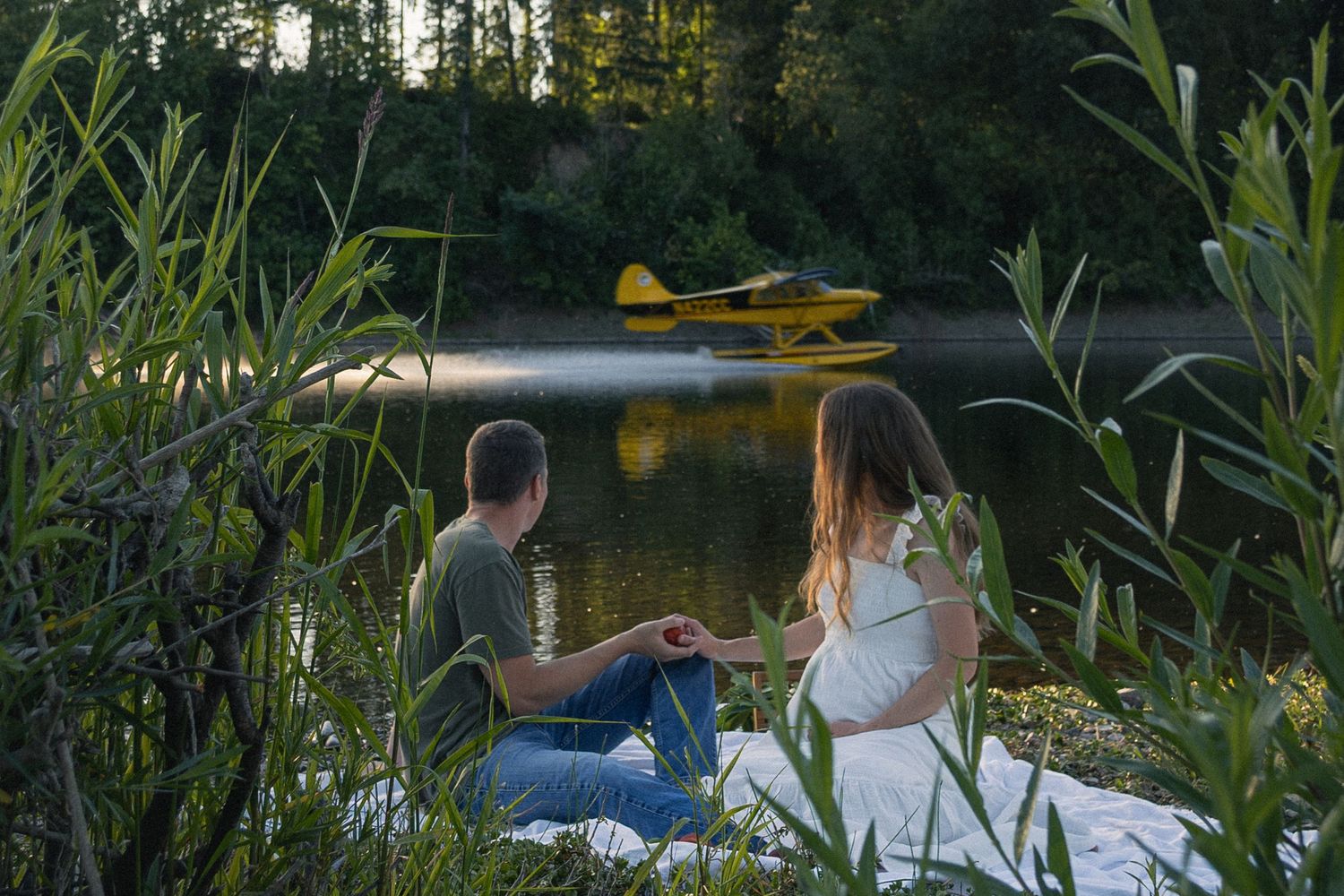 People enjoy a peaceful summer evening by a lake with a seaplane visible in the background.