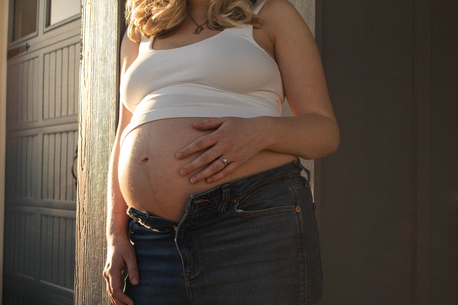 A pregnant person in a white tank top and jeans cradles their belly in soft natural light by a door.