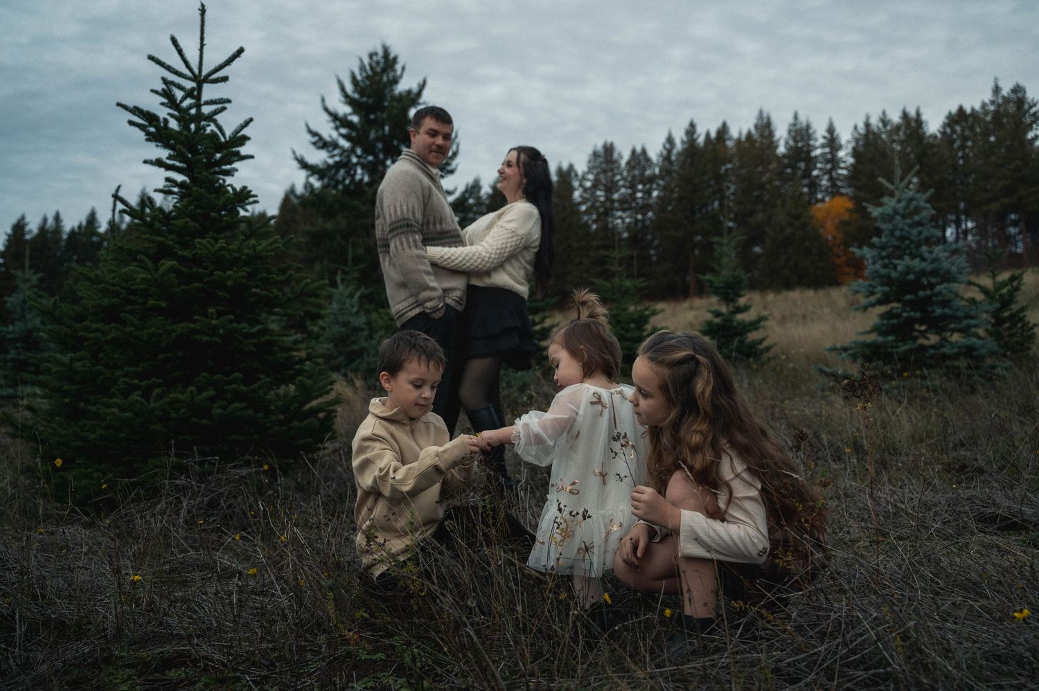 A group of people in winter coats embrace in a rustic outdoor setting with evergreen trees in the background.