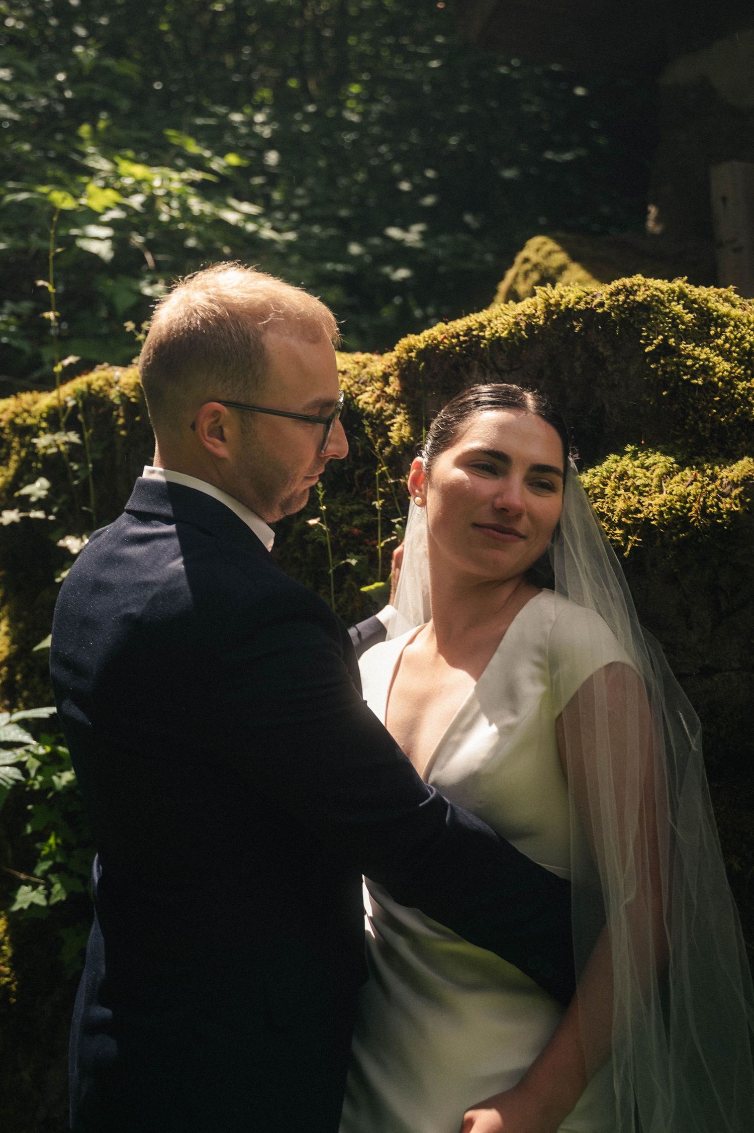 A bride in a white gown and veil shares a tender moment with her groom in a navy suit amid lush mossy greenery.