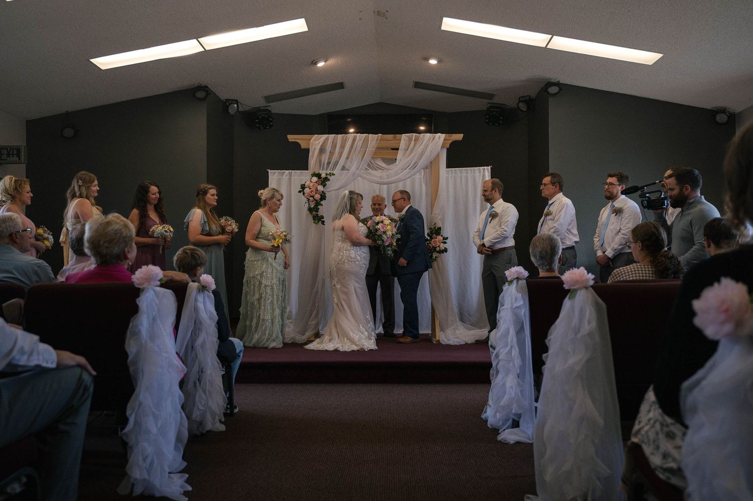 Wedding party members in formal attire gather in a dimly lit reception hall during a celebration.